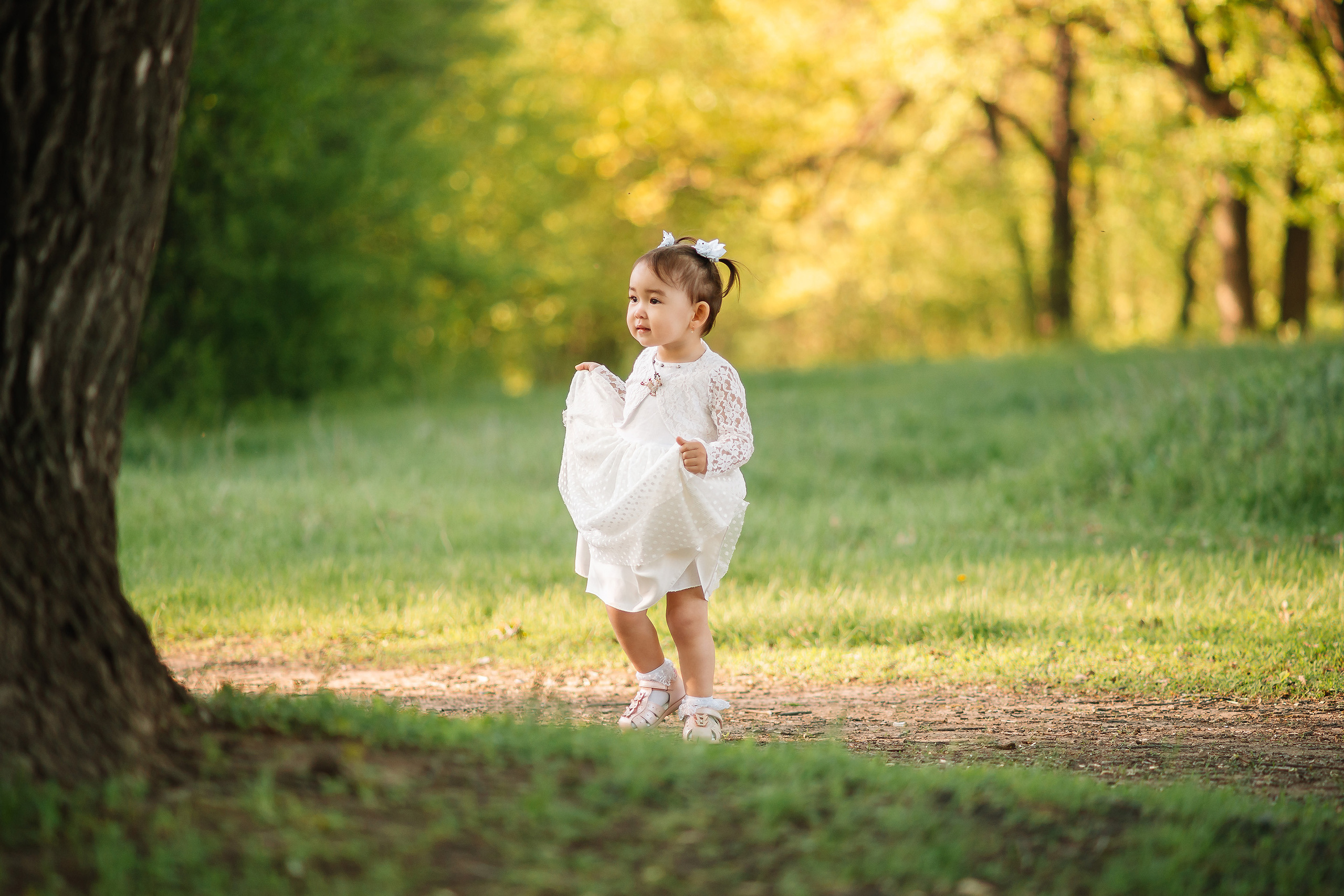 Маленькая казахская девочка в белоснежном воздушном платье посередине зеленой лужайки около большого старого дуба на фоне леса в лучах солнца смотрит куда-то в сторону.