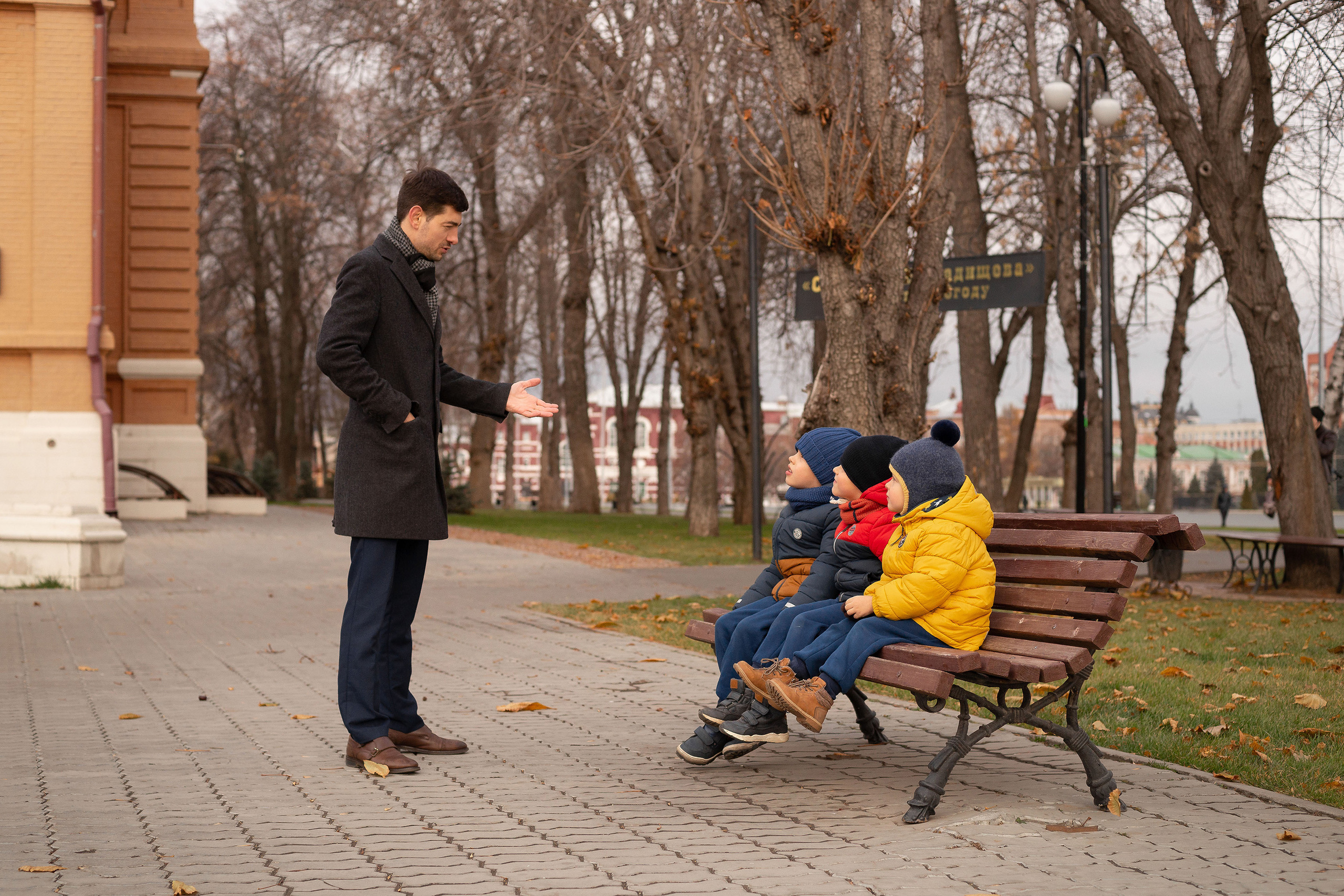 Трое братьев сидят на лавочке, а молодой отец стоит перед ними и что-то объясняет на фоне осенних деревьев.