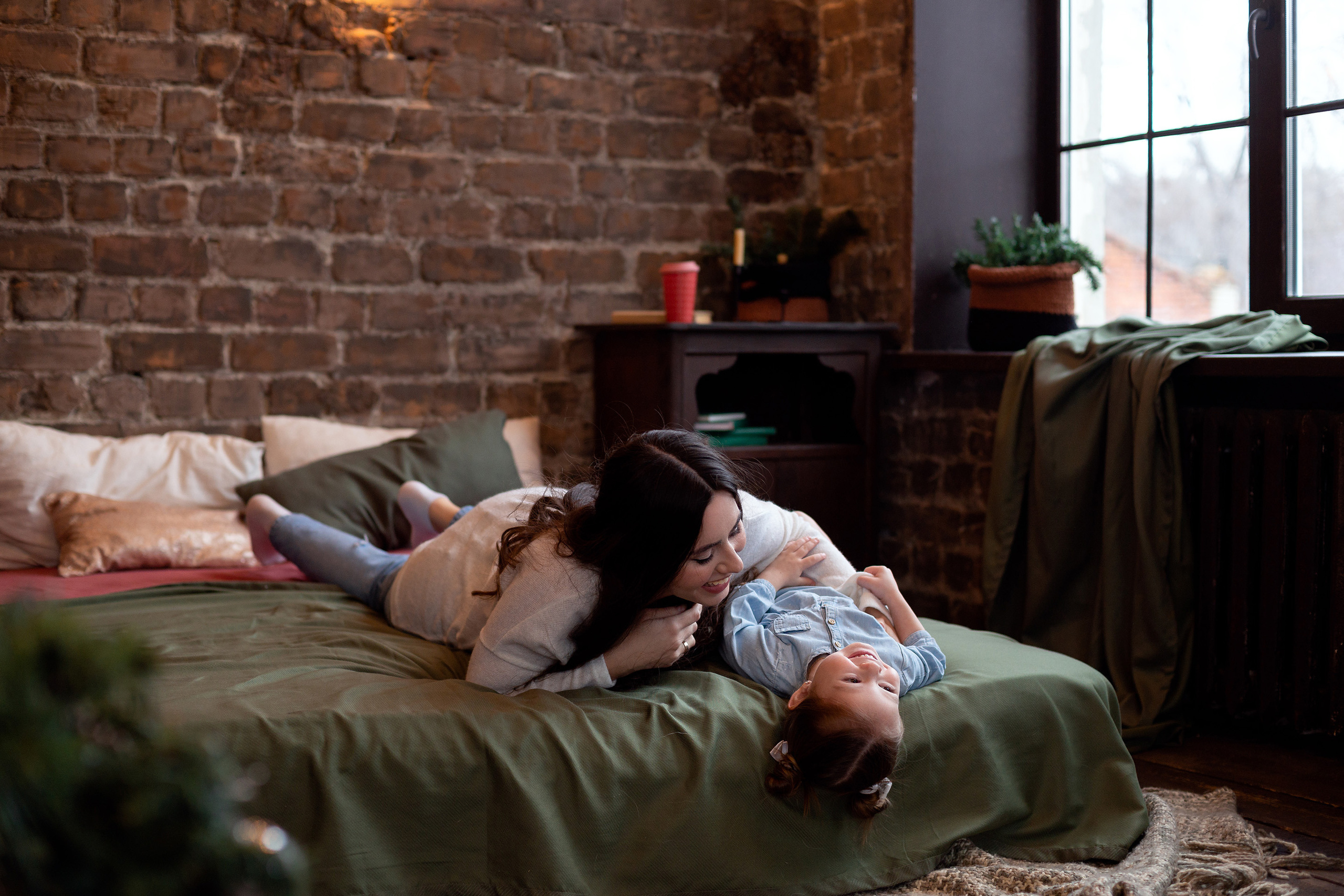 Мама щекочет маленькую дочку лежа на кровати в комнате в стиле лофт, и они счастливы.