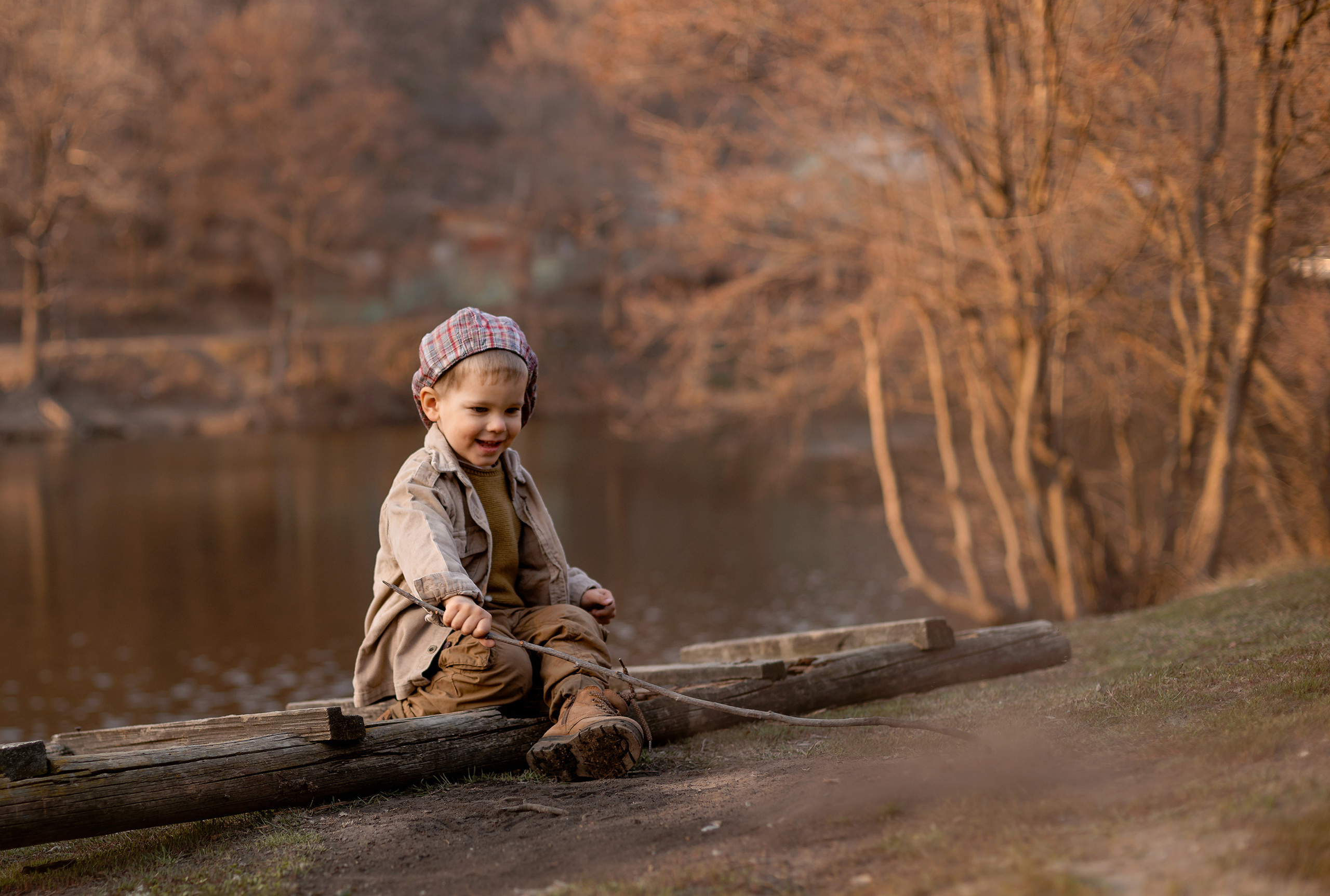Маленький мальчик сидит на бревне около пруда и играет с палкой.