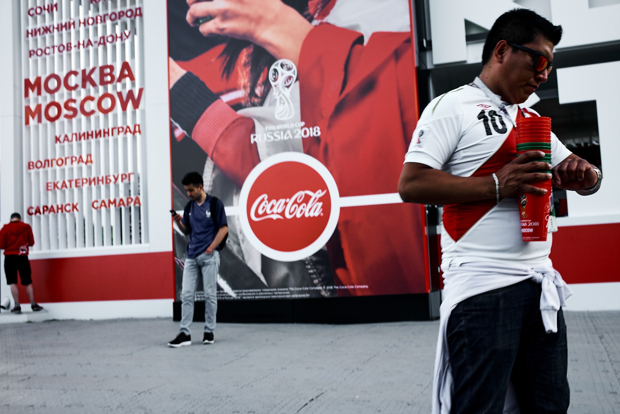 Болельщик с пластиковыми стаканами смотрит на часы на фоне рекламного баннера Coca-Cola и надписи Moscow Russia 2018. Жанровый портрет, решающий момент, уличная фотография. Фотограф Виктор Молодцов.