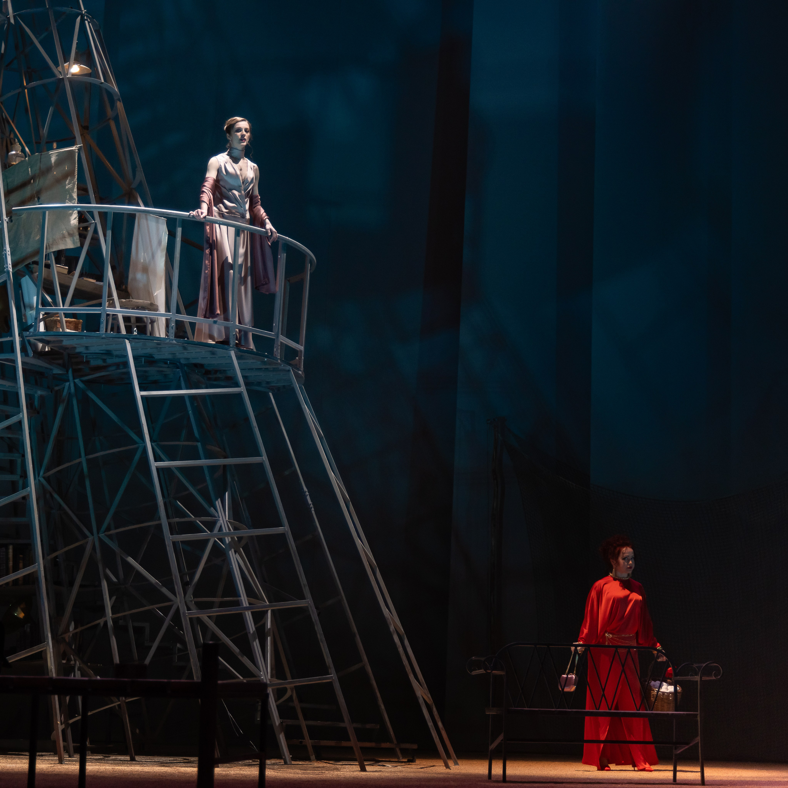 Женщина в светлом платье стоит на металлической лестнице, за ней темно-синяя сцена