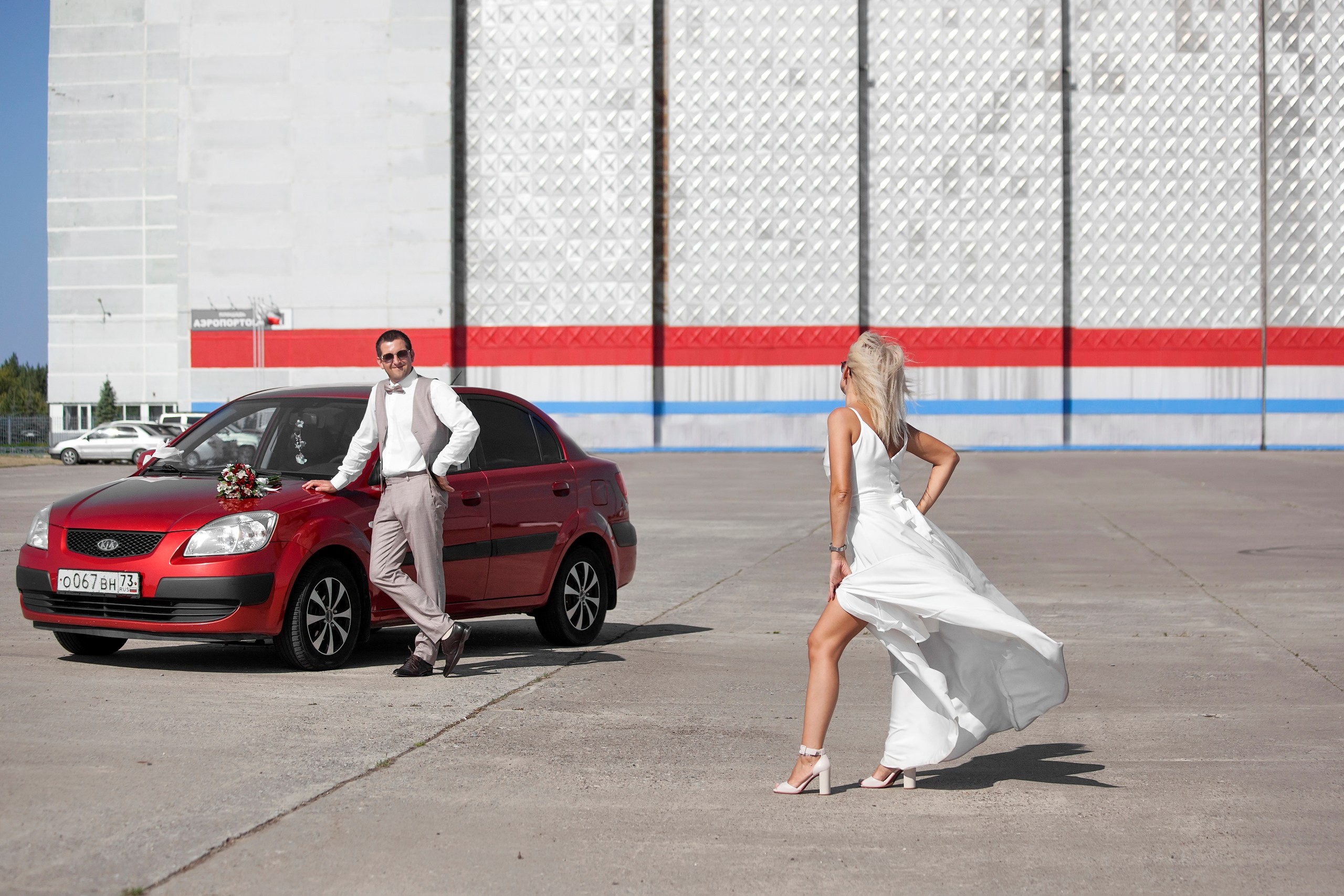 Невеста в развевающемся платье смотрит на жениха у красной машины, свадебная фотосъёмка