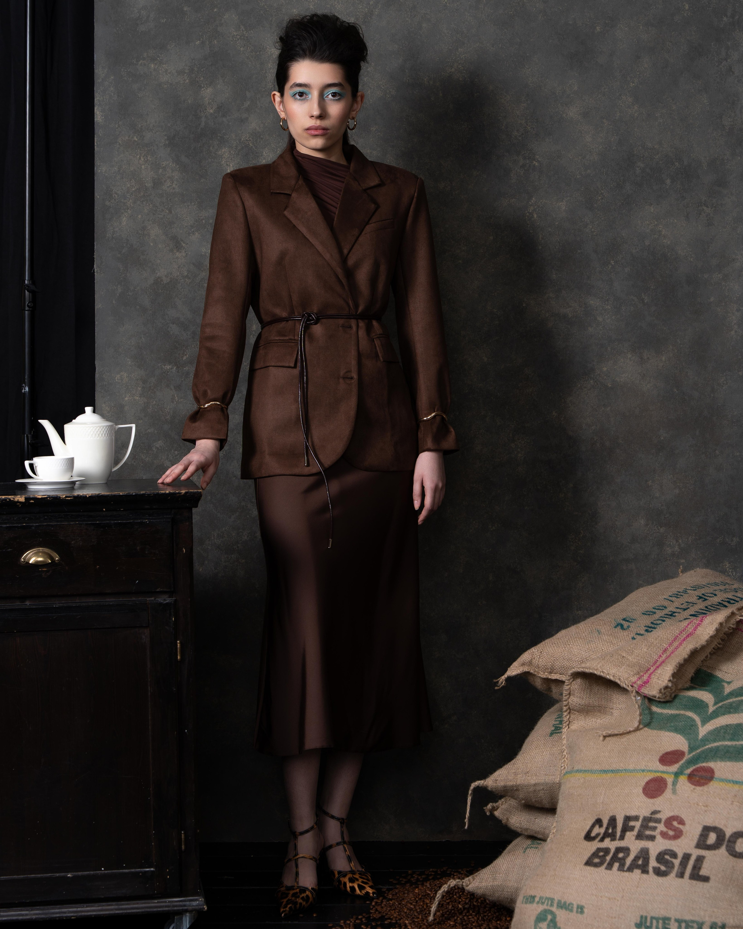 На фотографии изображена женщина, стоящая в интерьере, который напоминает кофейню или студию. Она одета в коричневый костюм, состоящий из пиджака и юбки, и носит черные туфли с леопардовым принтом. На заднем плане видны мешки с надписью "CAFÉ DO BRASIL", что указывает на бразильский кофе. На столе рядом с женщиной стоит белый чайник и чашка. Фон выполнен в темных тонах, что создает контраст с одеждой модели.