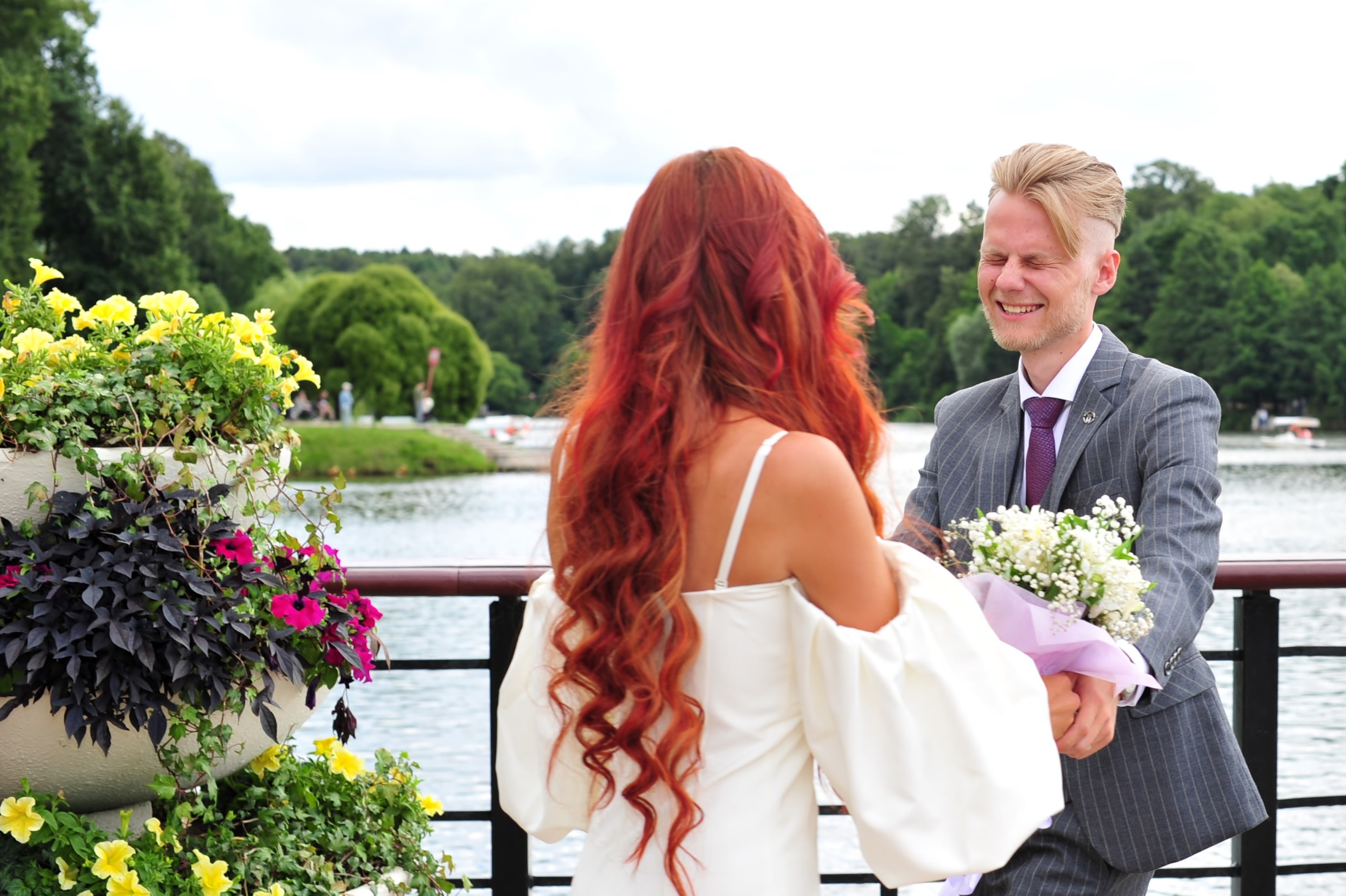 Свадьба в Царицыно летом, жених и невеста держатся на руки на фоне пруда, жених смеется