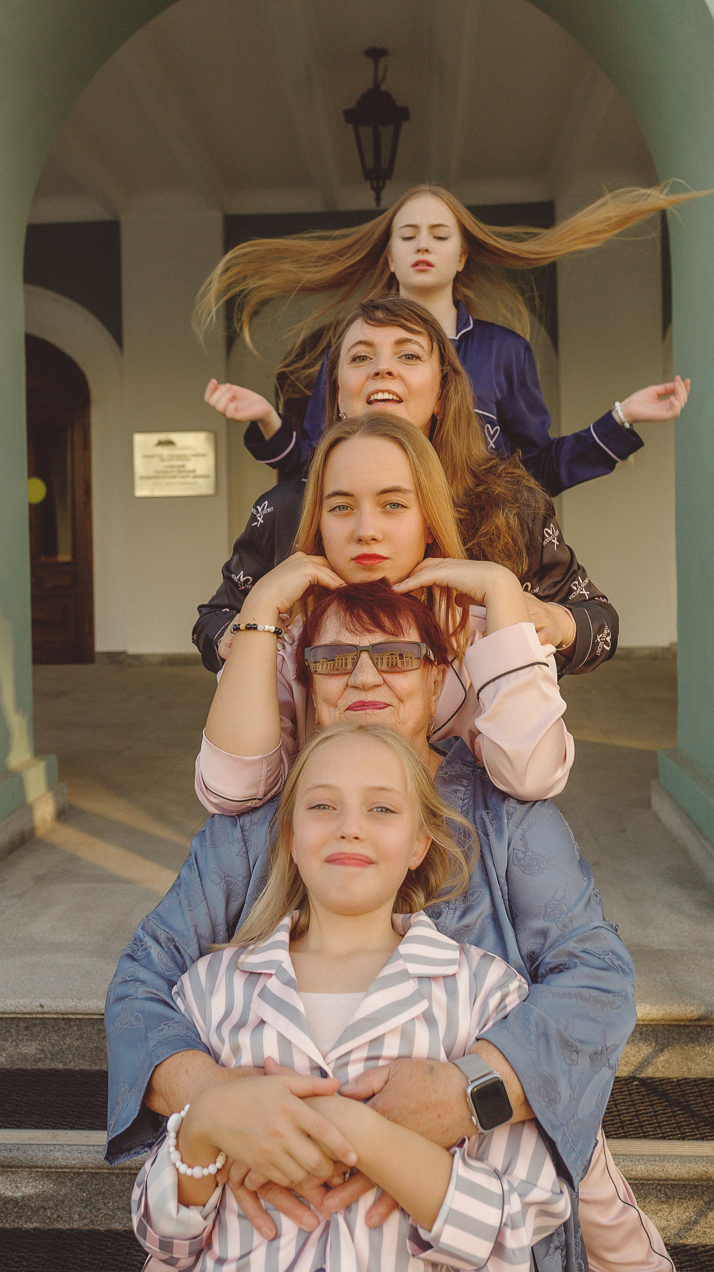 Вертикальная группа из пяти человек - бабушка, мама и три дочки - улыбается и игриво позирует перед драмтеатром. Они передают ощущение радости и единения.