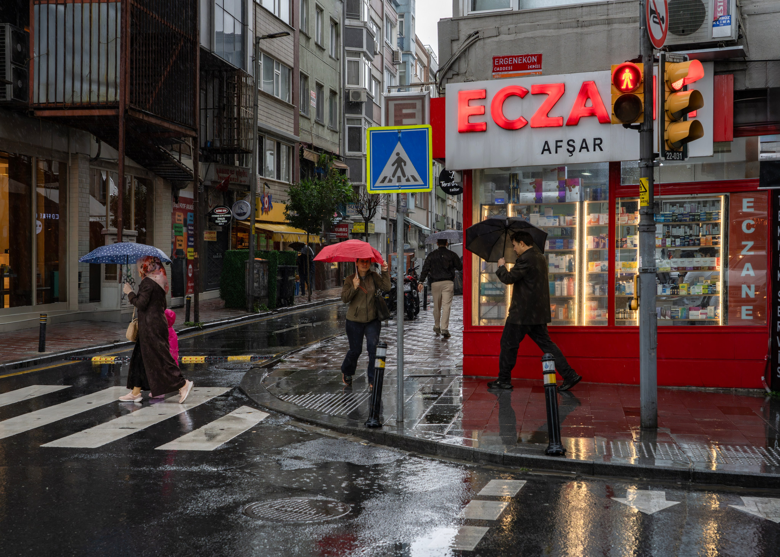 Urban street photography of rainy Istanbul, people with umbrellas at a crosswalk near a pharmacy building with a red ECZANE sign.