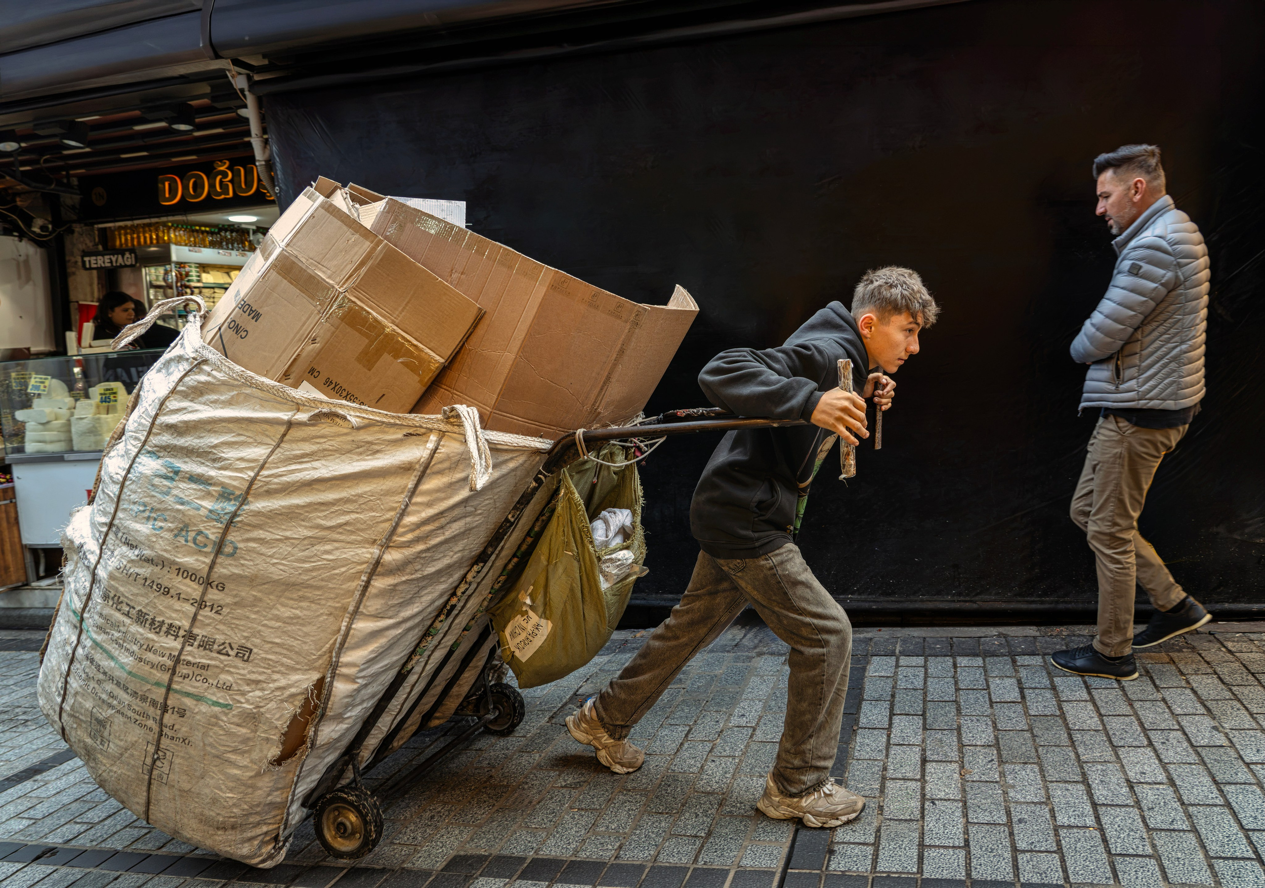 A young waste paper collector (çekçekçi) pulls an overloaded cart with cardboard boxes along a street in Istanbul.