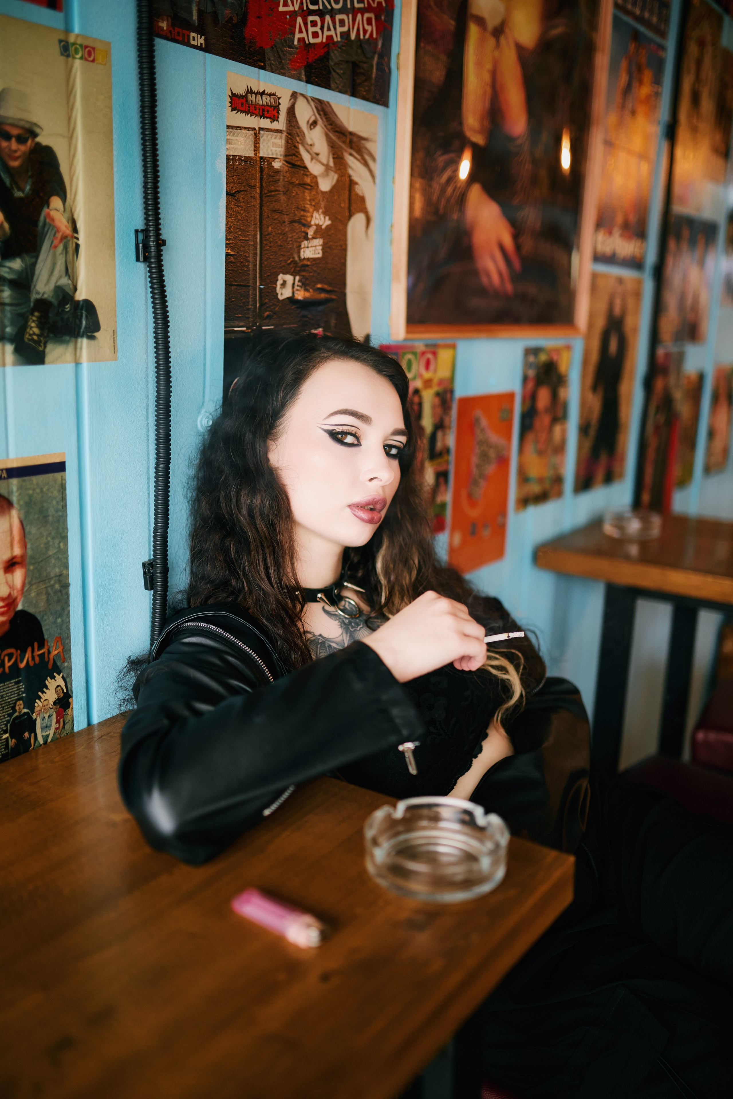 Портрет курящей девушки в кожаной куртке, позирующей в баре на фоне плакатов рок-групп
