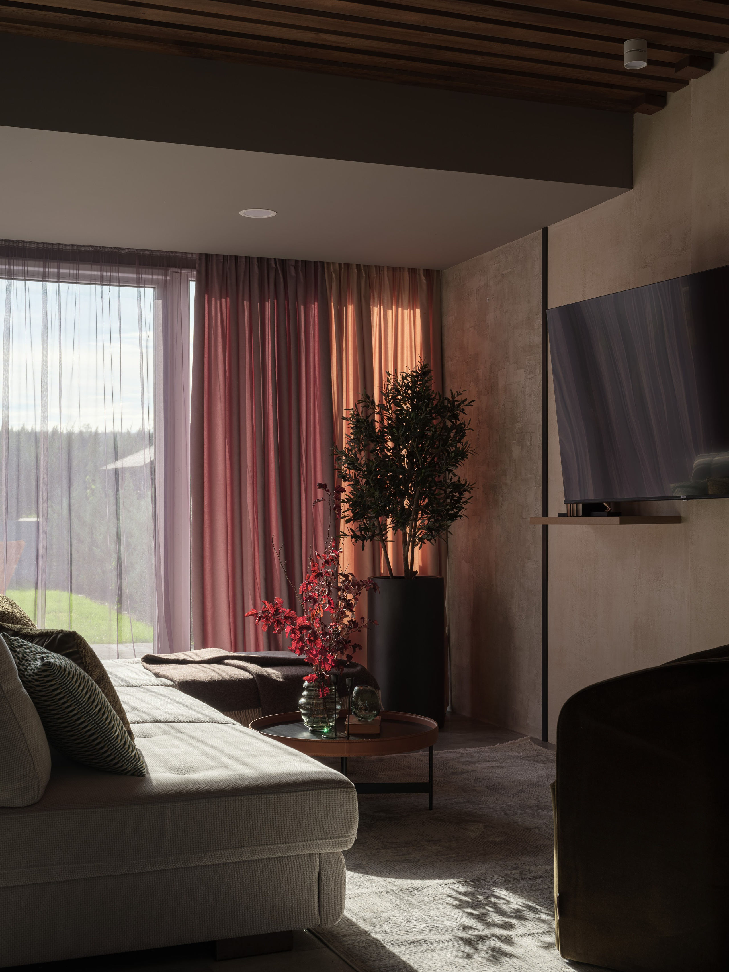 Современная гостиная с панорамными окнами, мягким диваном и акцентными шторами оттенков терракоты и розы. Декор с живыми растениями и вазы с алыми ветвями создают уют и теплоту. Интерьер оформлен дизайнером Алиной Дёровой, стилист Наталья Якушева, фото Сергей Денисенко.