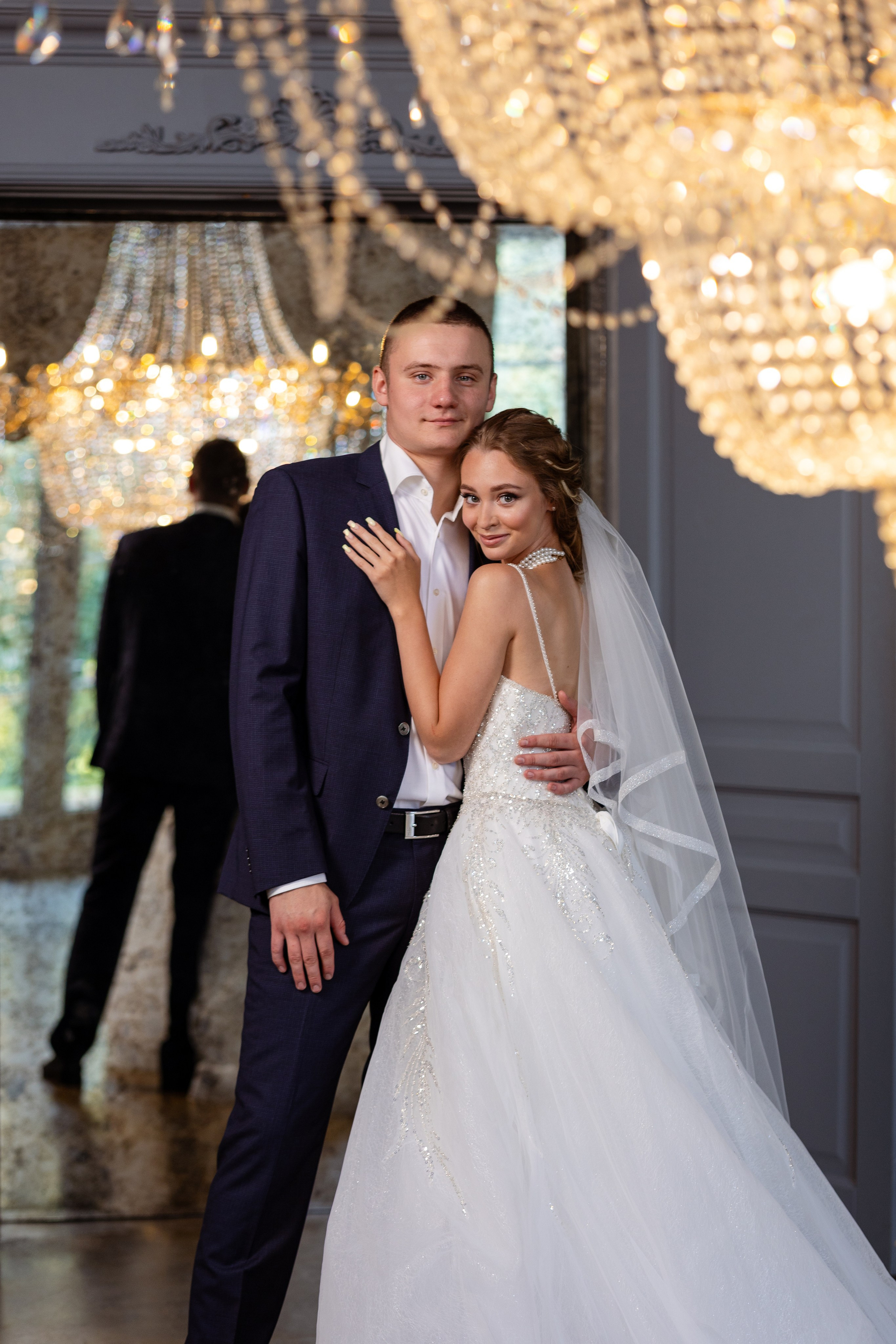 Молодые стоят в обнимку на фоне двух огромных сияющих люстр.