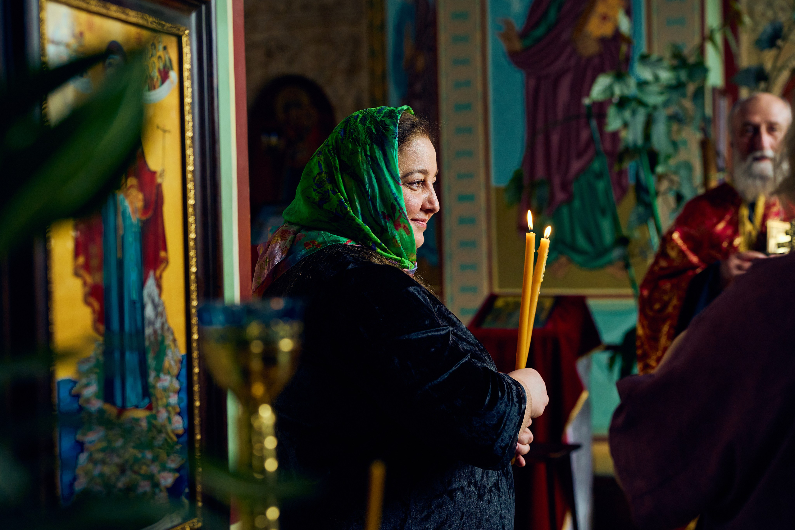 Женщина в зелёном платке держит свечи напротив иконы в православной церкви, за ней священник