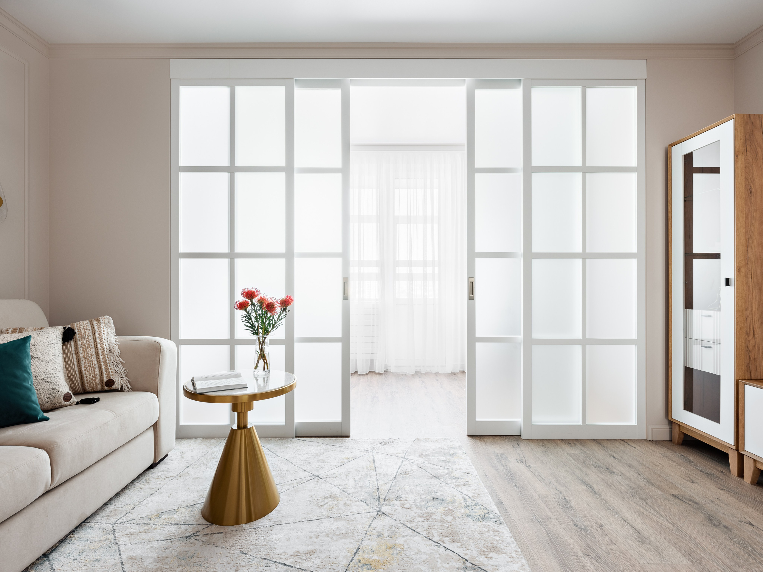 Панорамный вид гостиной с раздвижными белыми дверями с матовым стеклом, современным ковром, мягким диваном и стильным золотым столиком с вазой. Гармоничную композицию создали дизайнер Наталья Бунеева, стилист Наталья Якушева, фотограф Сергей Денисенко.