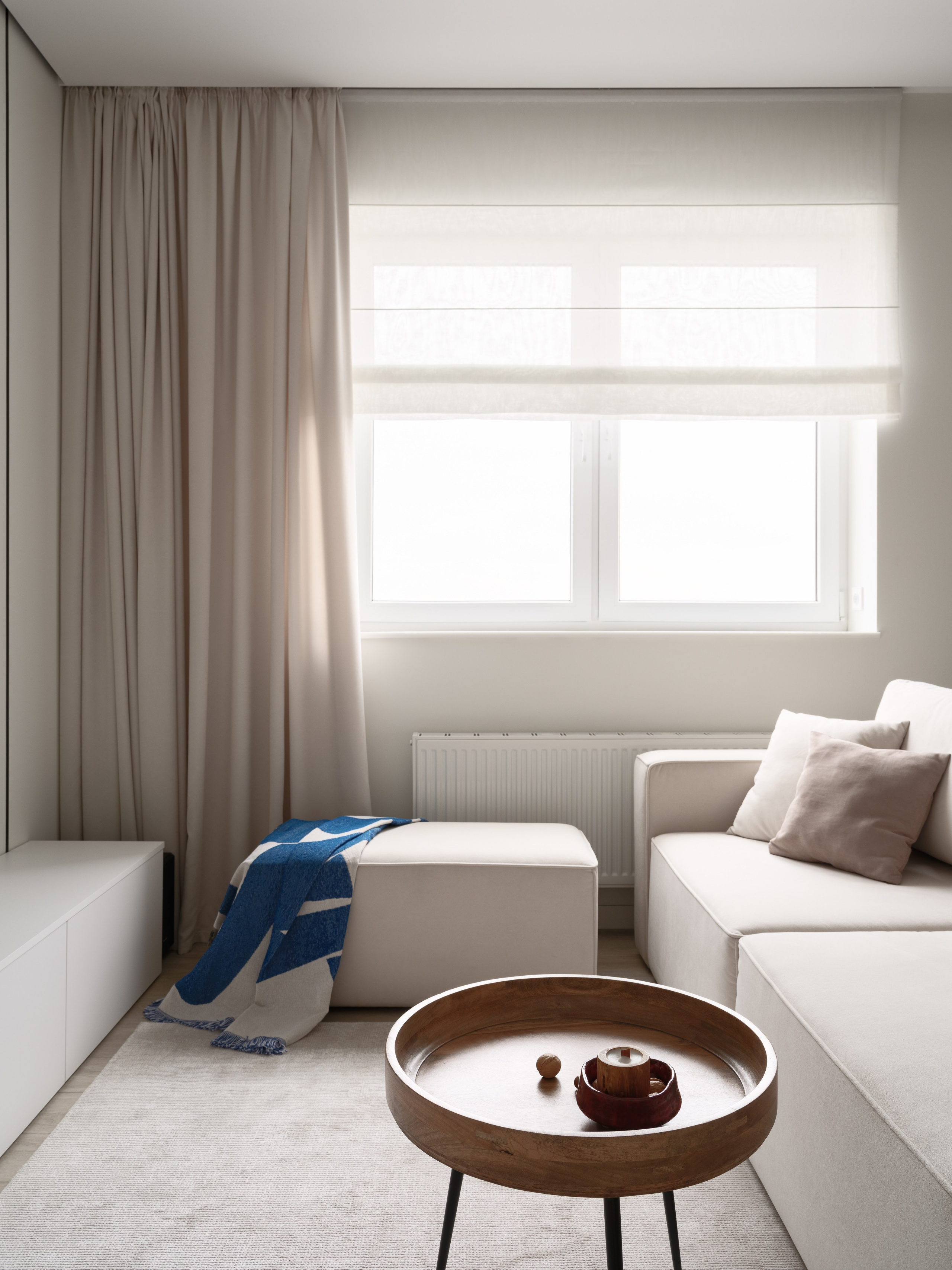 Современная гостиная с бежевым диваном, римскими шторами и акцентным синим пледом на пуфе. Минималистичный интерьер от дизайнеров Евгении Княжевой и Владлены Приваловой. Фотограф — Сергей Денисенко