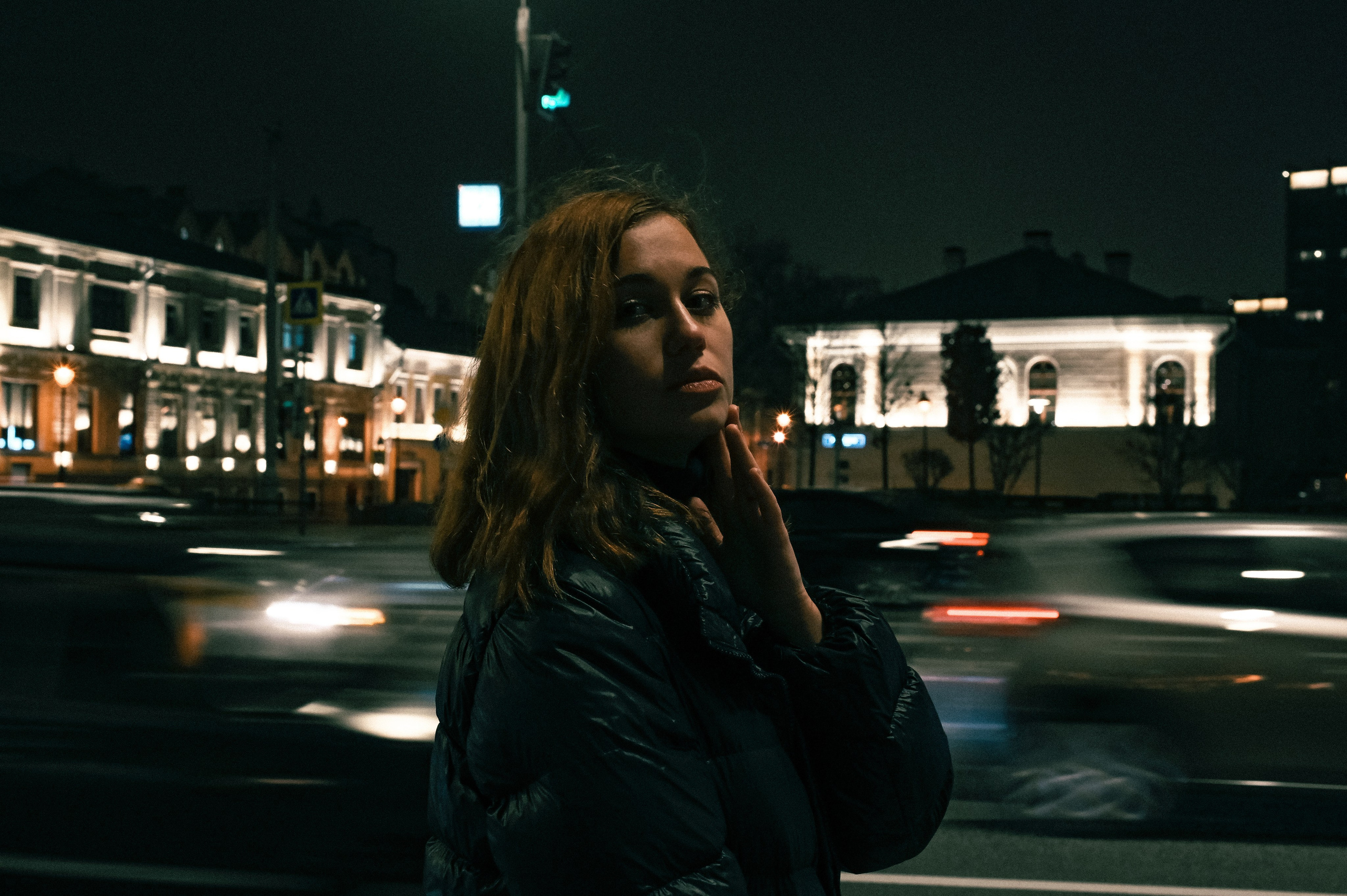 Ночной портрет девушки с волнистыми волосами в Москве, снятый у дороги с эффектом движения автомобилей и архитектурным фоном.