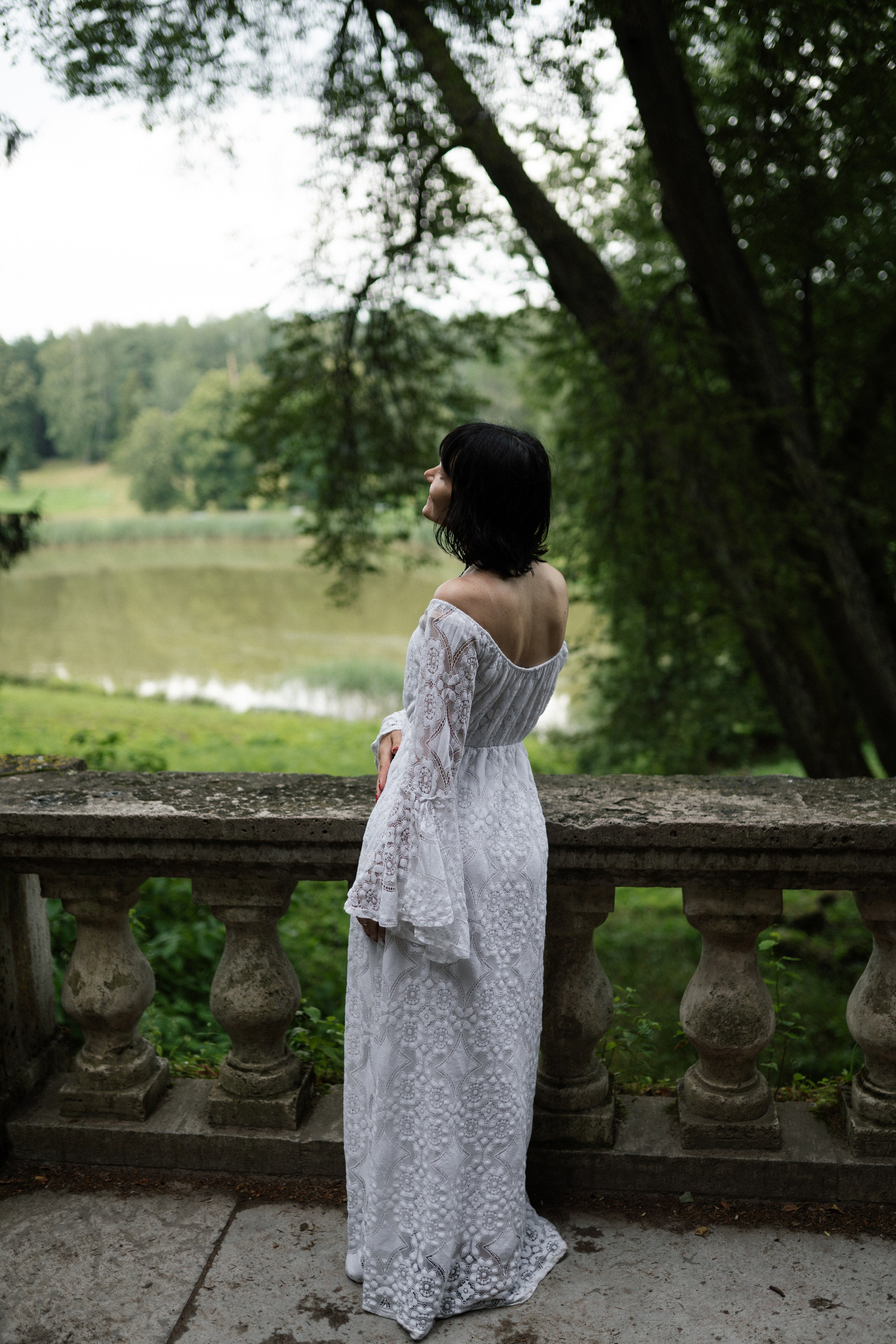 Модель в длинном белом кружевном платье стоит у каменного балкона у озера, художественная портретная фотосессия на природе в Санкт-Петербурге