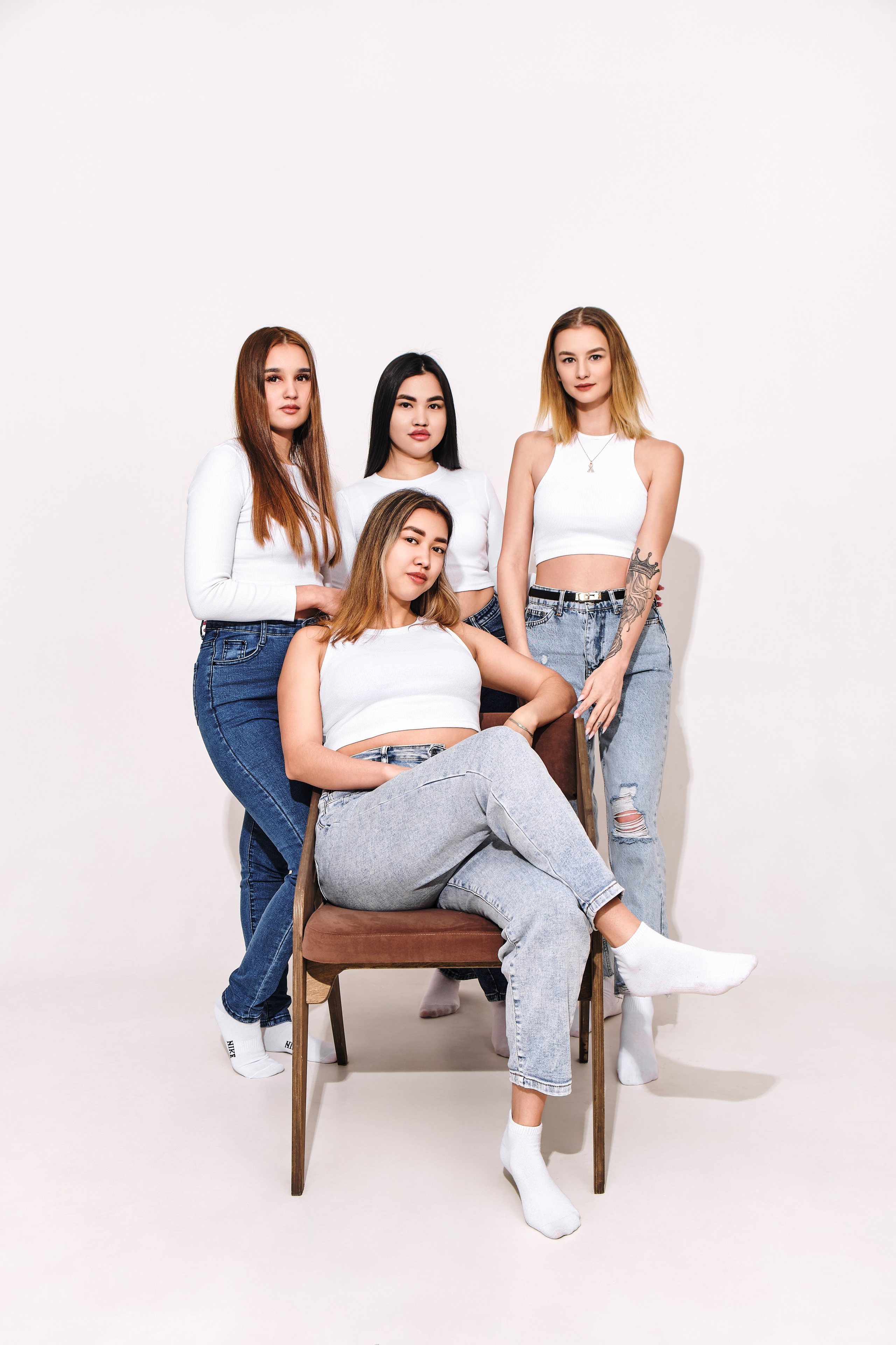 Групповой портрет четырех лучших подруг в одинаковых белых футболках и джинсах, искренне смеющихся на светло-сером фоне.