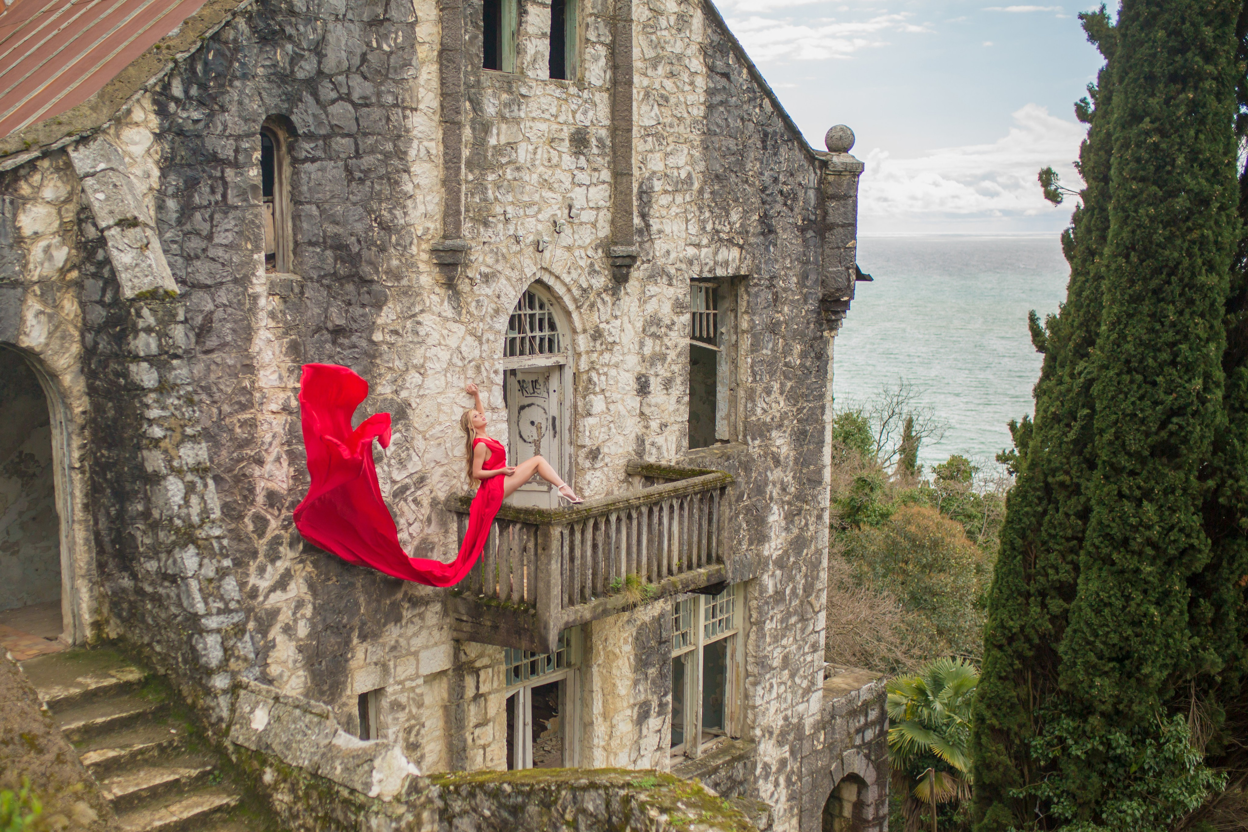 Девушка сидит в красном платье со шлефом на балконе заброшенного здания