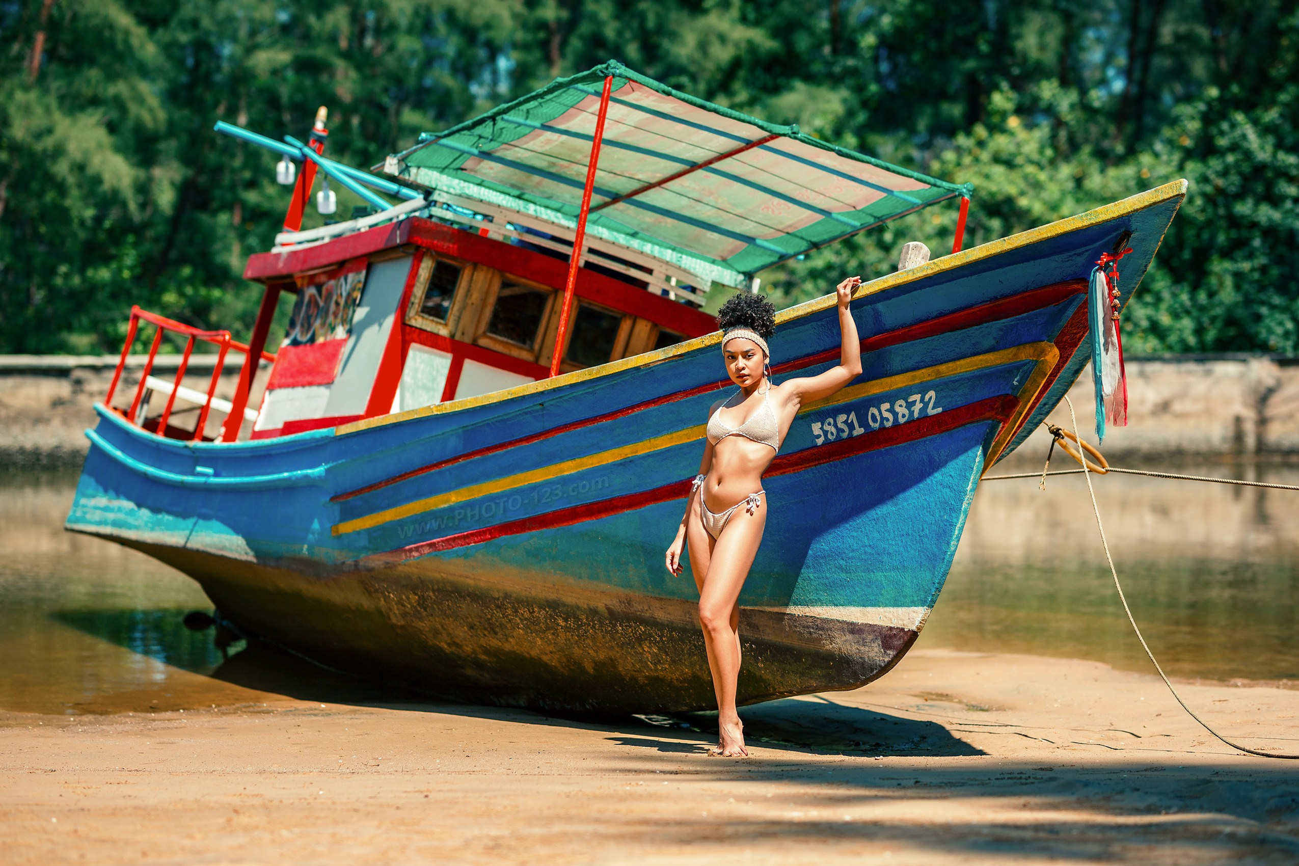 Confident woman in bikini posing beside colorful traditional fishing boat on sandy shore, tropical travel lifestyle, exotic beach photography with wooden boat and calm river