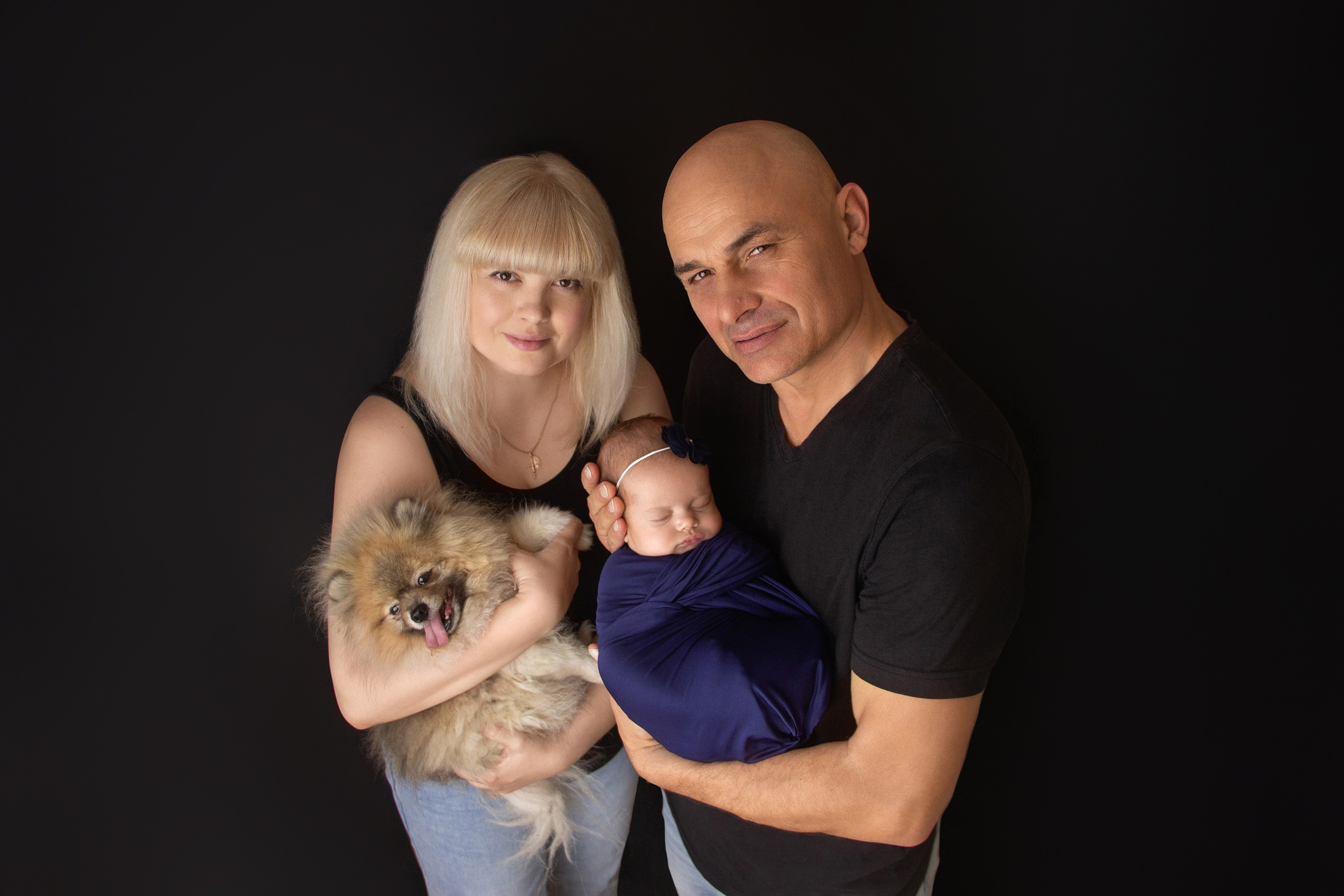 семейный кадр с молодыми красивыми родителями с собакой и новорожденной в обмотке стоят счастливые на фоне черной стены в Краснодаре 