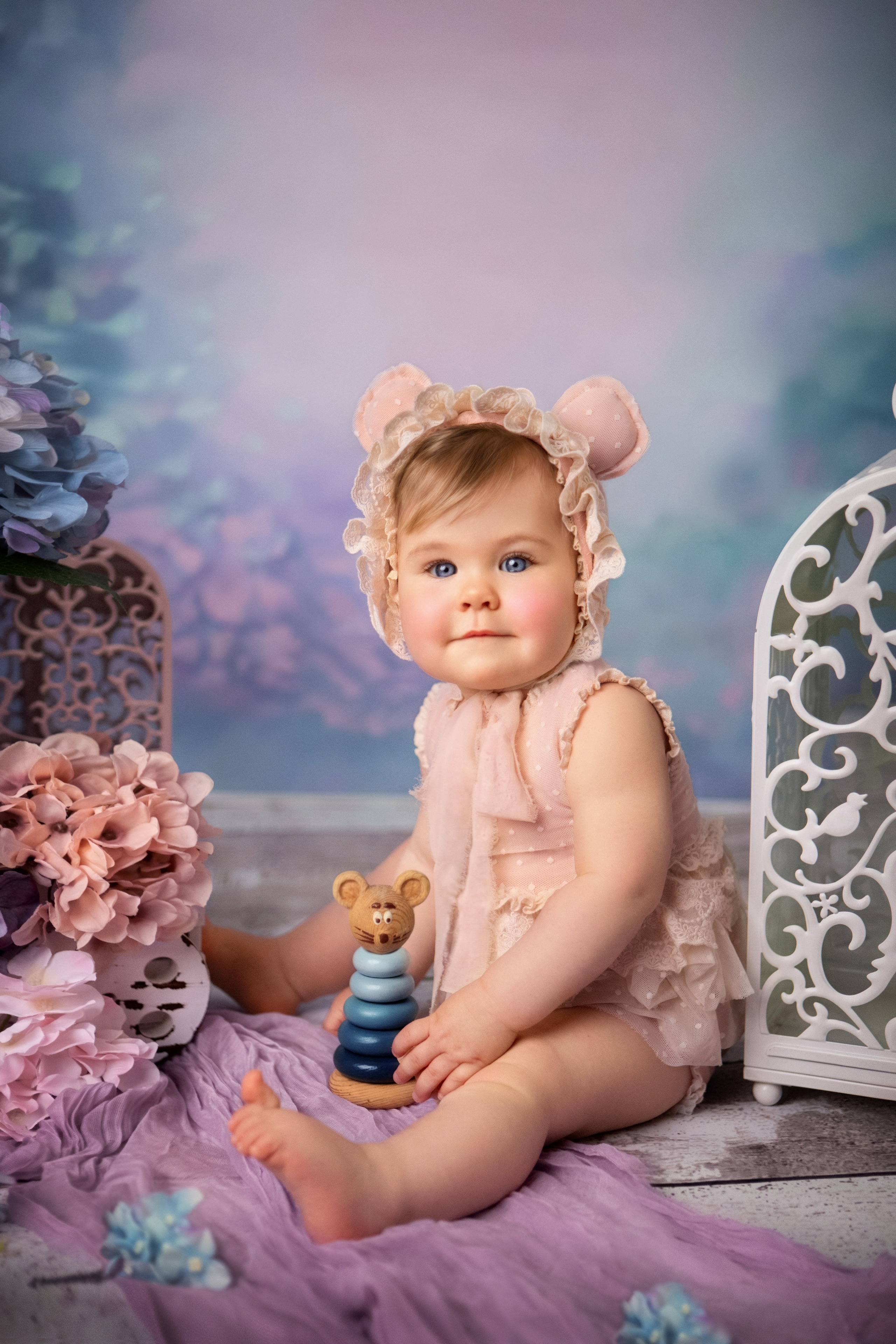 малышка в кружевном наряде ручной работы с ушками медвежонка сидит рядом с деревянной пирамидкой. Вокруг нее - цветы гортензии.