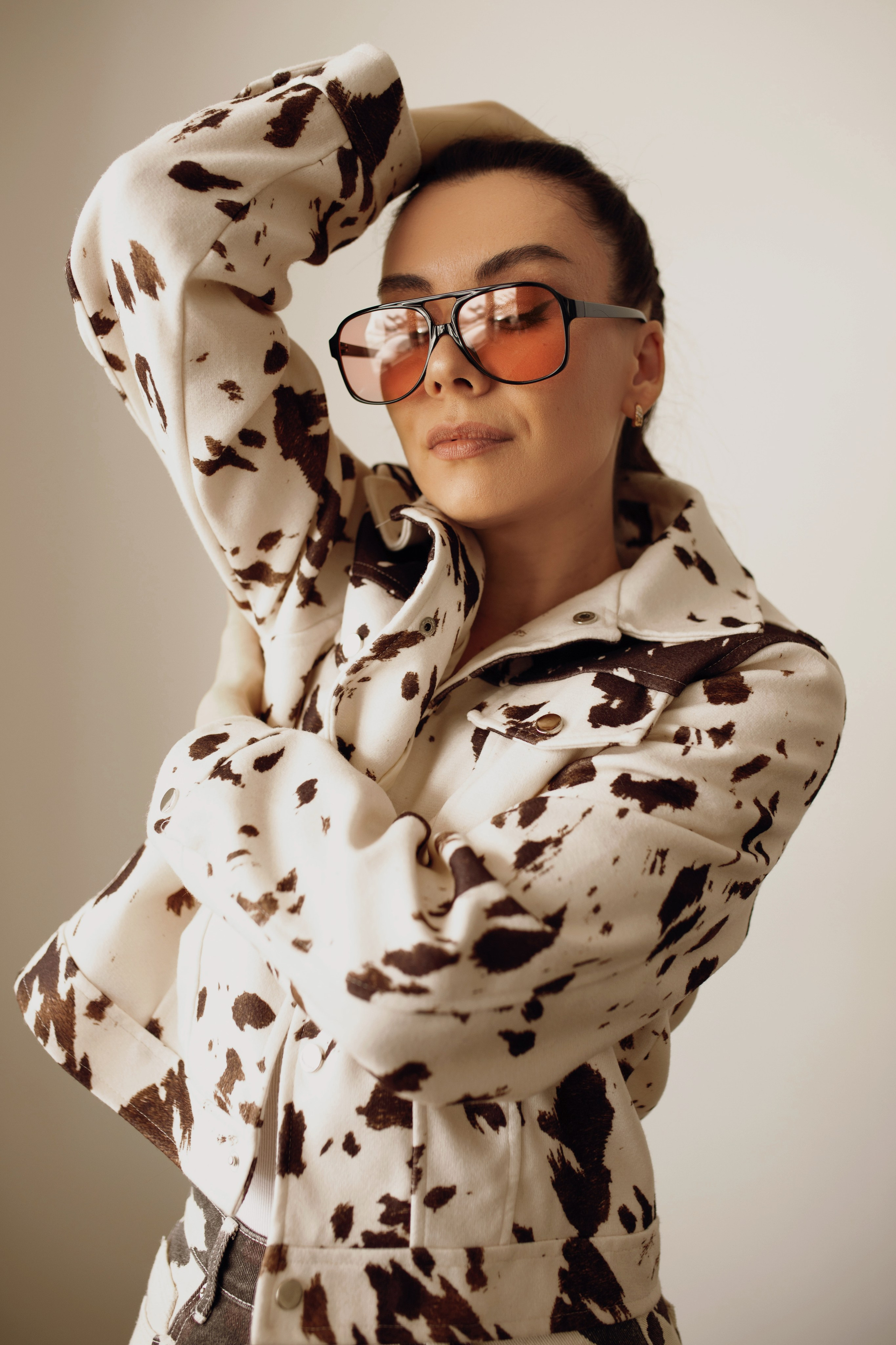 Женщина в стильной куртке с принтом коровы, в больших очках с оранжевыми линзами, позирует, подняв руку к голове.