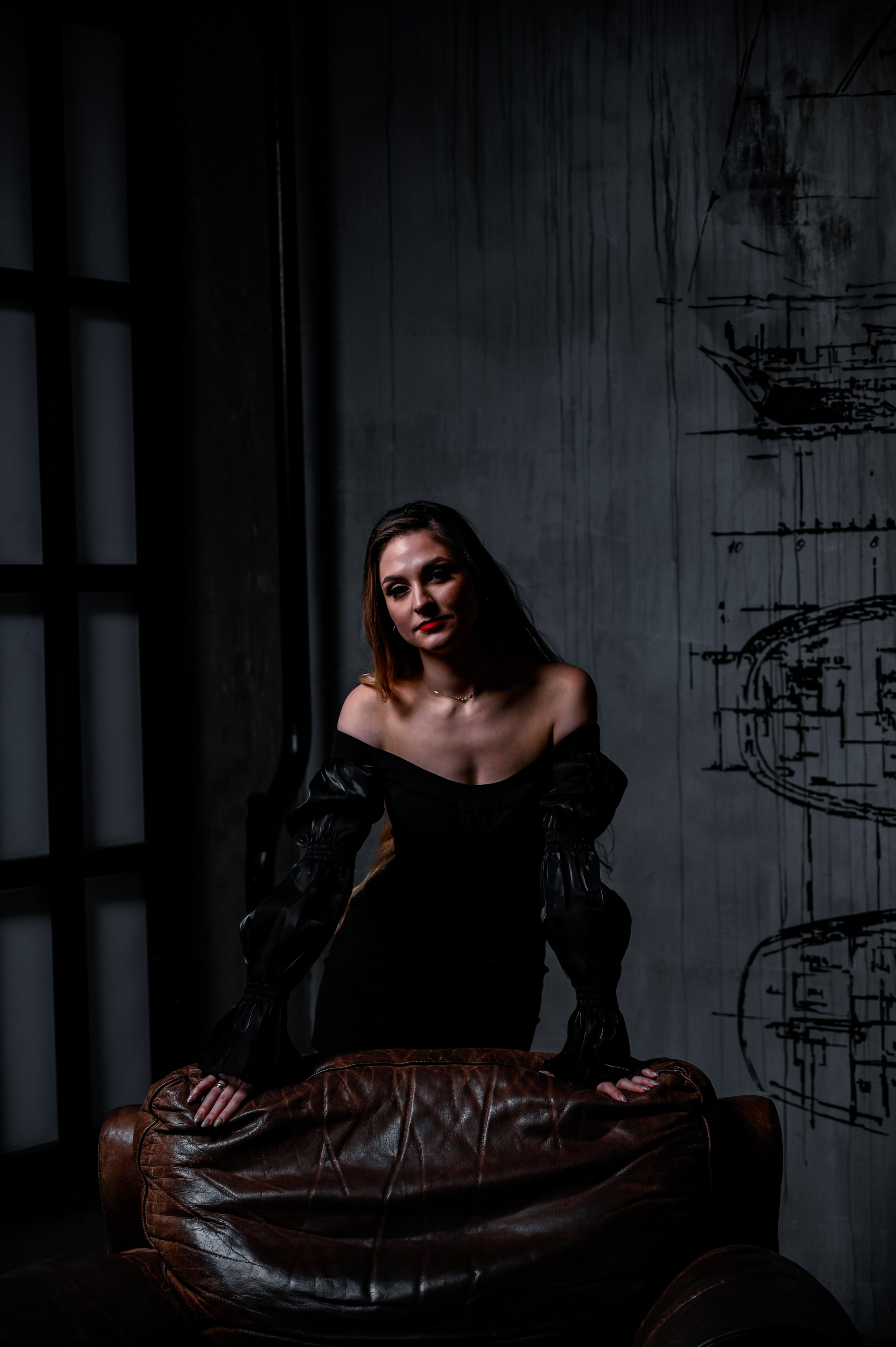 Молодая женщина сидит на кожаном диване в атмосферной, темно-тонированной комнате. Она одета в черное платье с открытыми плечами и длинными рукавами, которое подчеркивает её фигуру и создает элегантный, загадочный образ. Её волосы свободно падают по плечам, а взгляд направлен прямо в камеру, демонстрируя уверенность и сосредоточенность. На лице у нее акцентный макияж с ярко-красными губами, который контрастирует с тёмным фоном. Вокруг модели видны элементы интерьера, такие как окно со ставнями и графические рисунки на стене, что добавляют атмосферу таинственности и интриги. Фотография передает чувственную и стильную атмосферу.