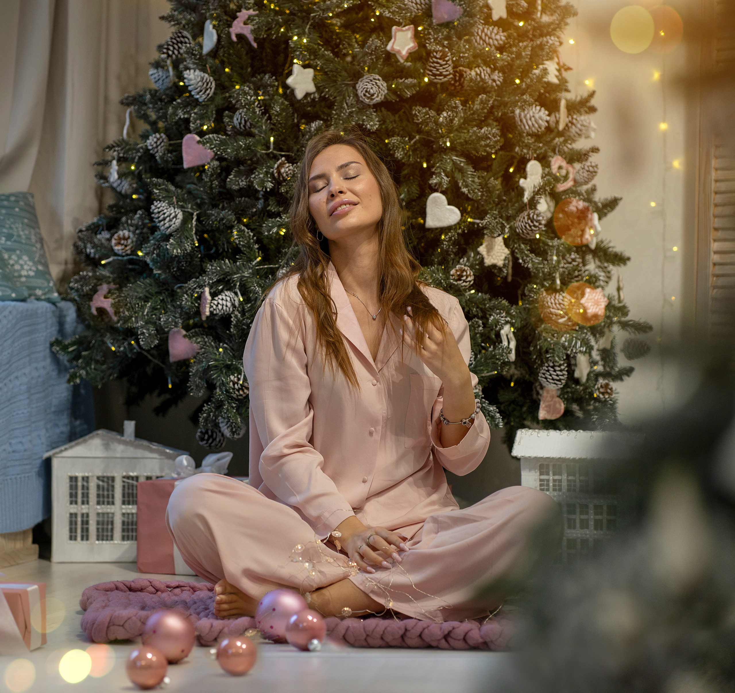 Девушка в пижаме сидит у новогодней елки, мечтает о подарках.