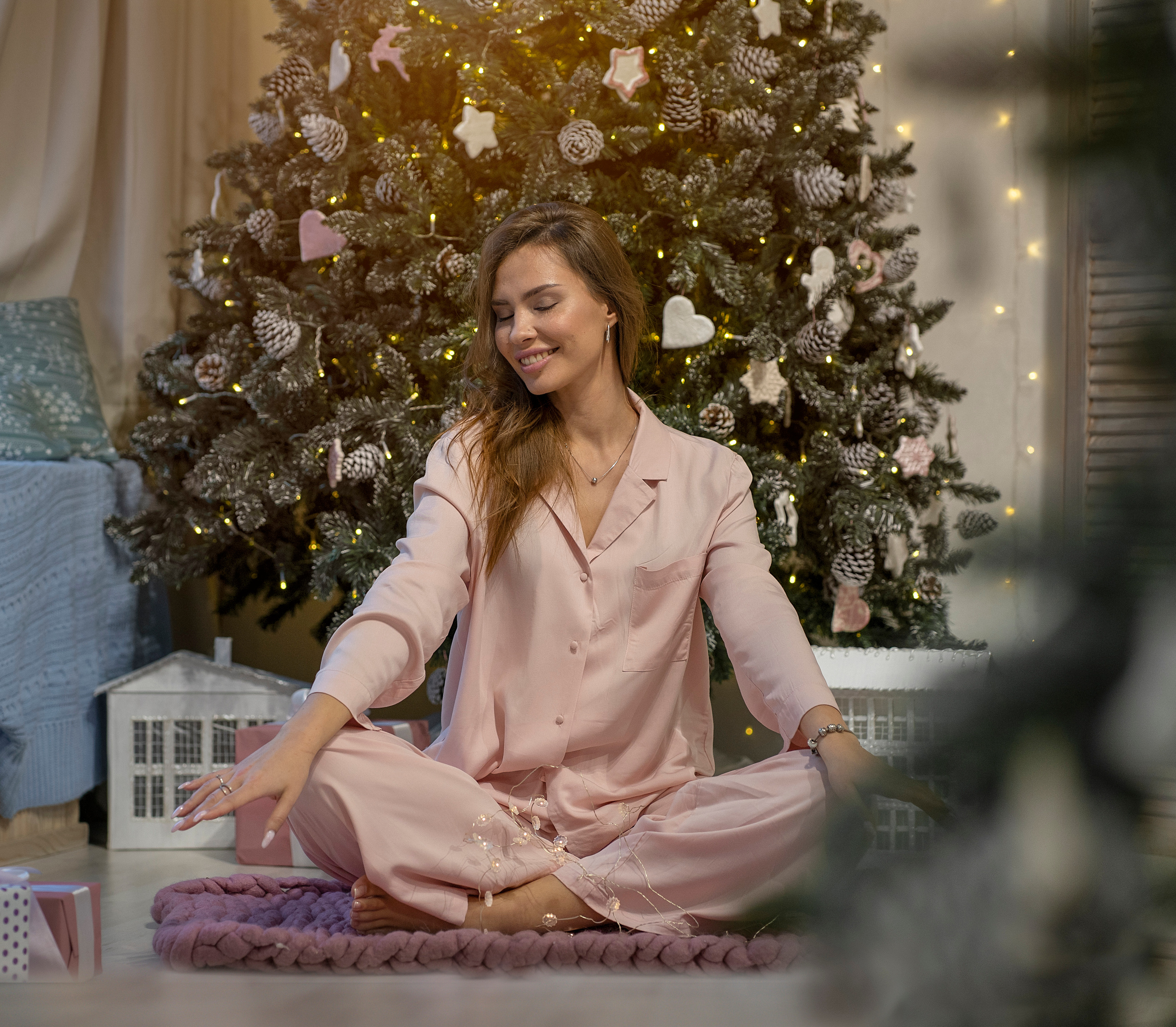Комната по новогоднему украшена. Девушка в пижаме сидит на полу, её весело.