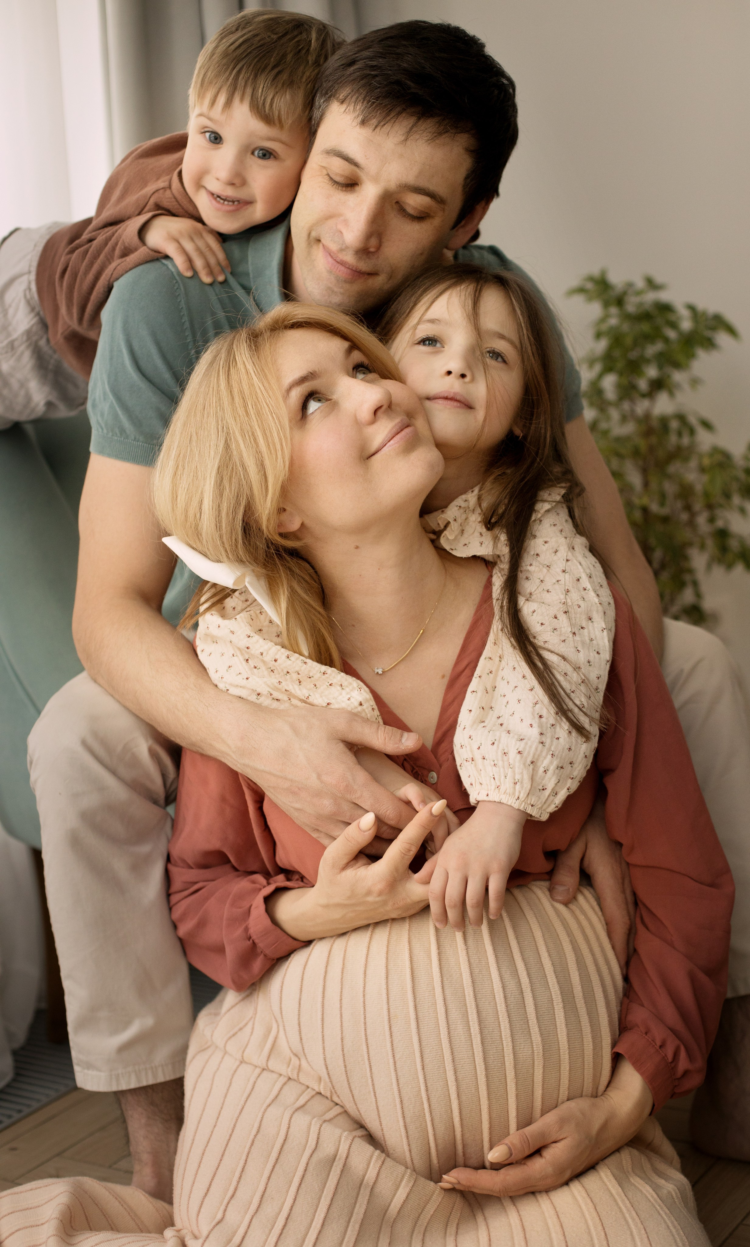 Беременная мама, папа, дочка и сын обнимаются сидя у окна на кресле