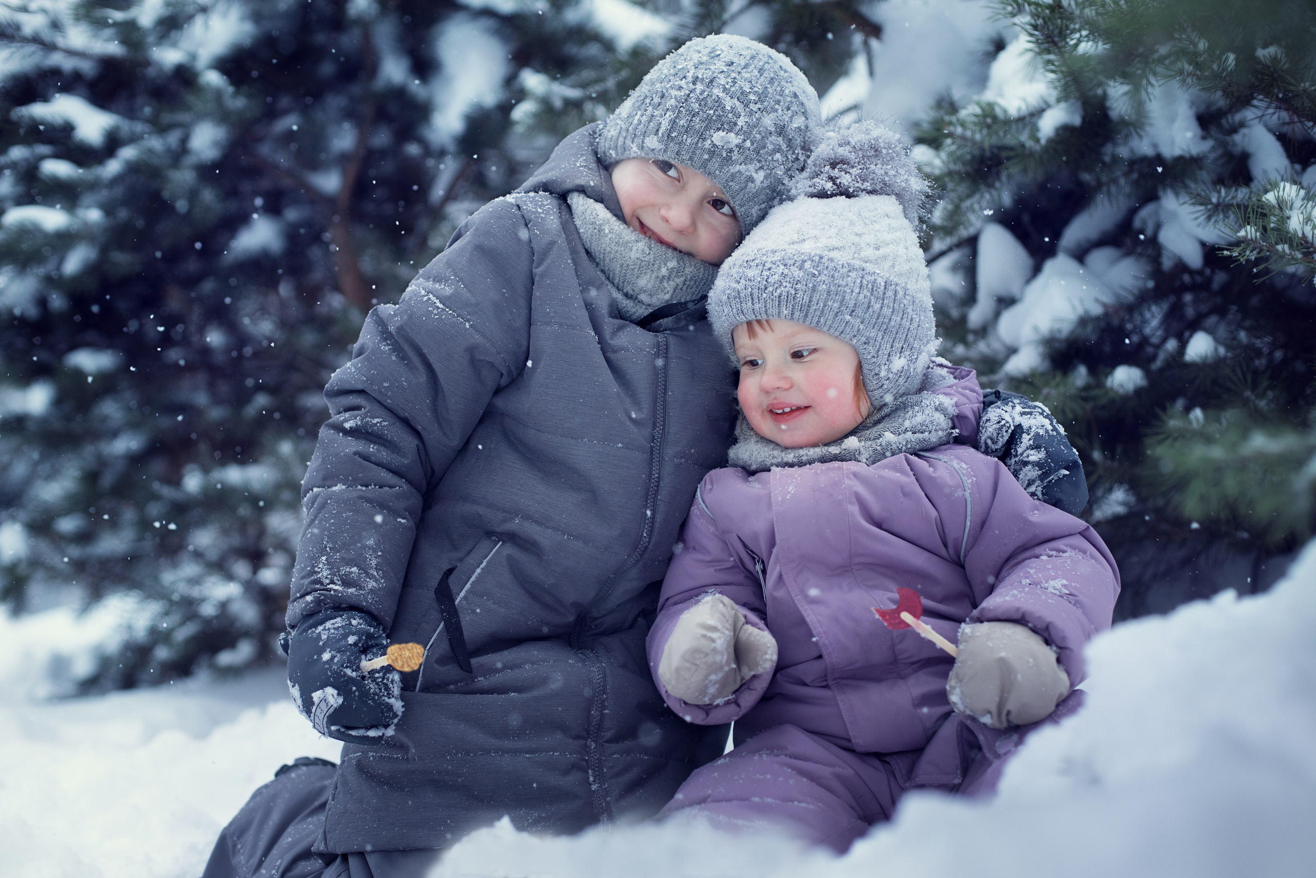 Девочка и мальчик сидят на снегу в зимнем лесу с леденцами в руках