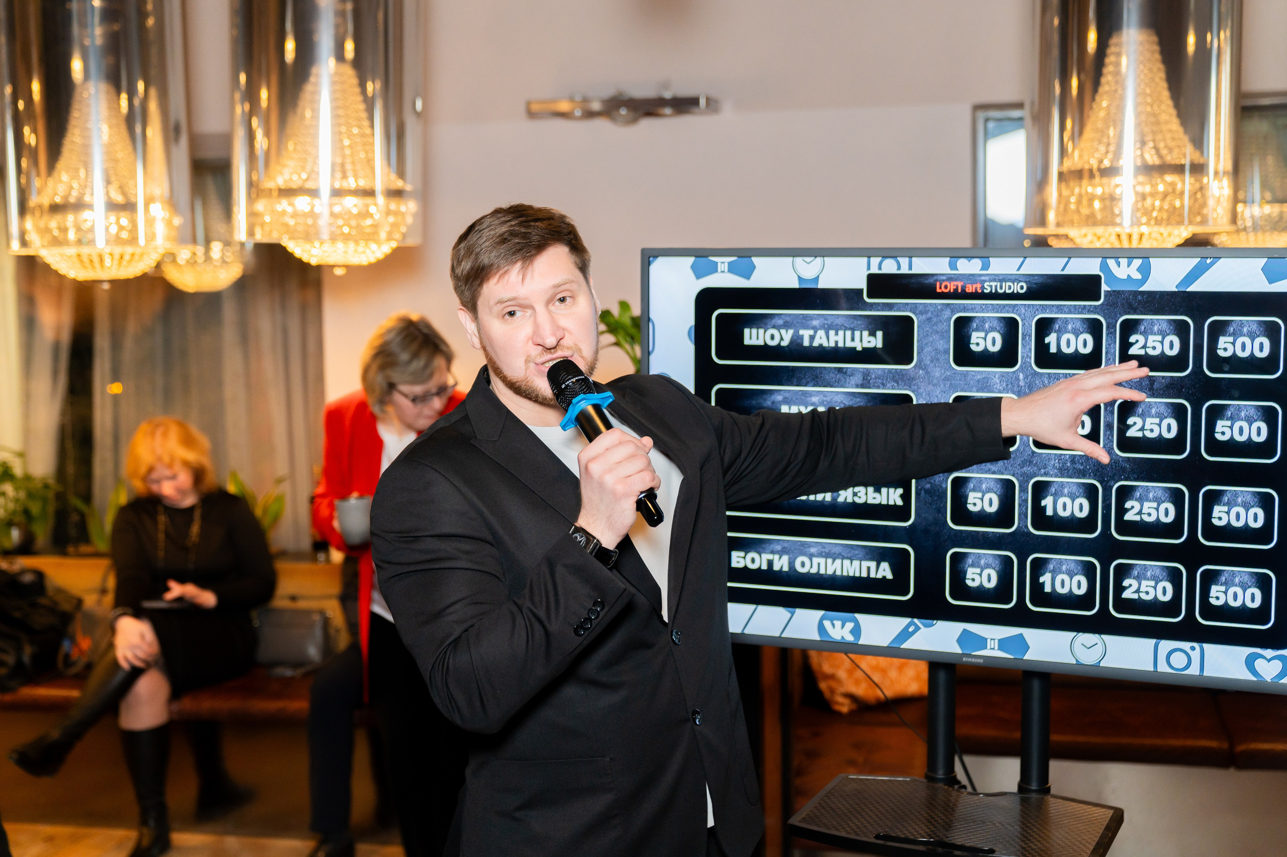 Ведущий в чёрном пиджаке с микрофоном показывает игровое табло на большом экране во время мероприятия в Radisson Sonya. Фотография Евгения Мальцева (evrednix.ru).