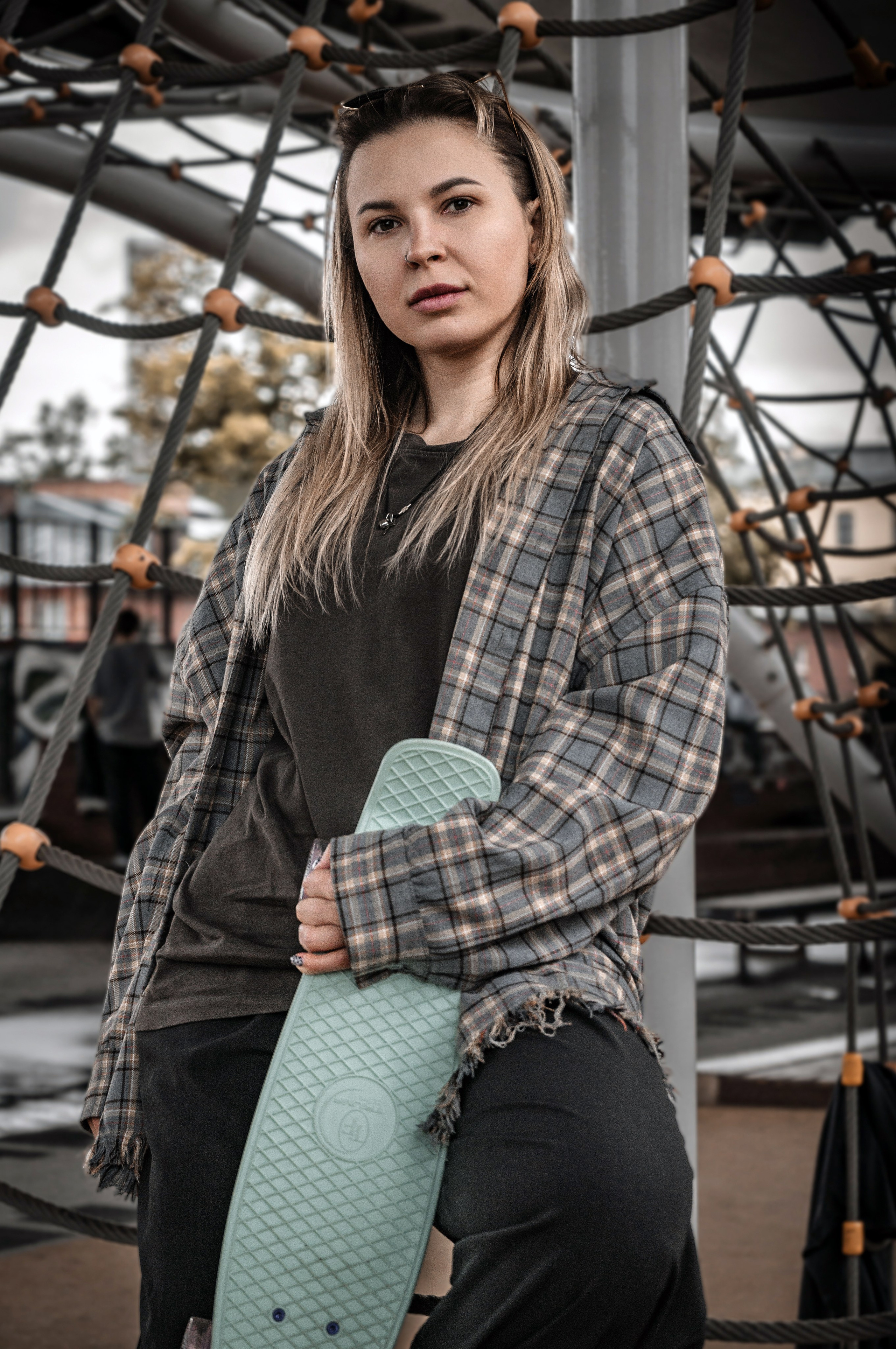 Стиль streetwear: девушка с пирсингом стоит на детской площадке с мятным penny board в руках, oversize клетчатая рубашка в клетку, широкие чёрные брюки, расслабленная поза, урбанистическая скейт-атмосфера.