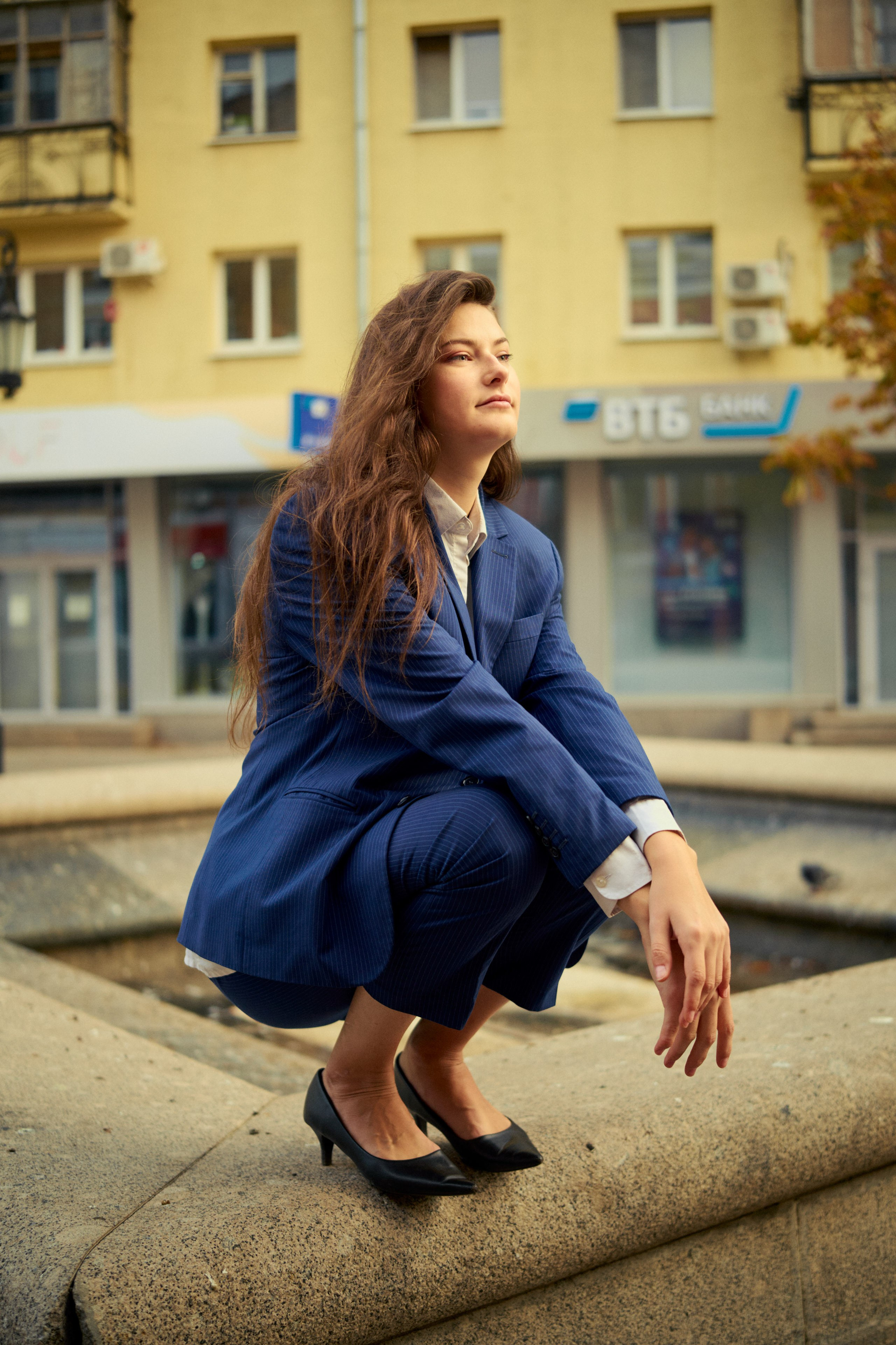  Уверенная девушка в классическом костюме, сидящая рядом с городским фонтаном на фоне архитектурного ландшафта