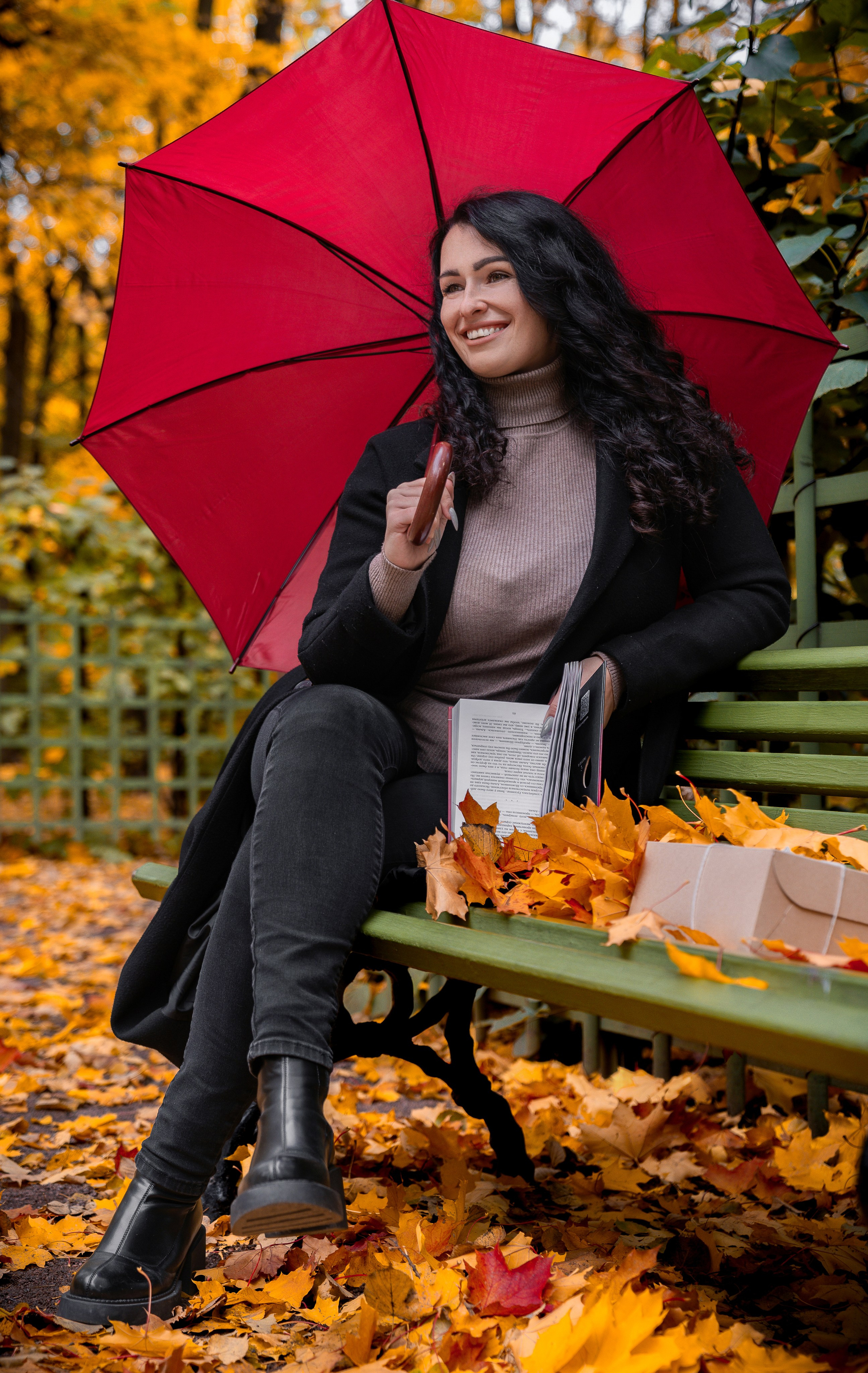 Красивая брюнетка в чёрном пальто и сером свитере сидит на скамейке в осеннем парке с красным зонтом и книгой, осенняя прогулочная фотосессия СПб, фотограф Евгений Мальцев.