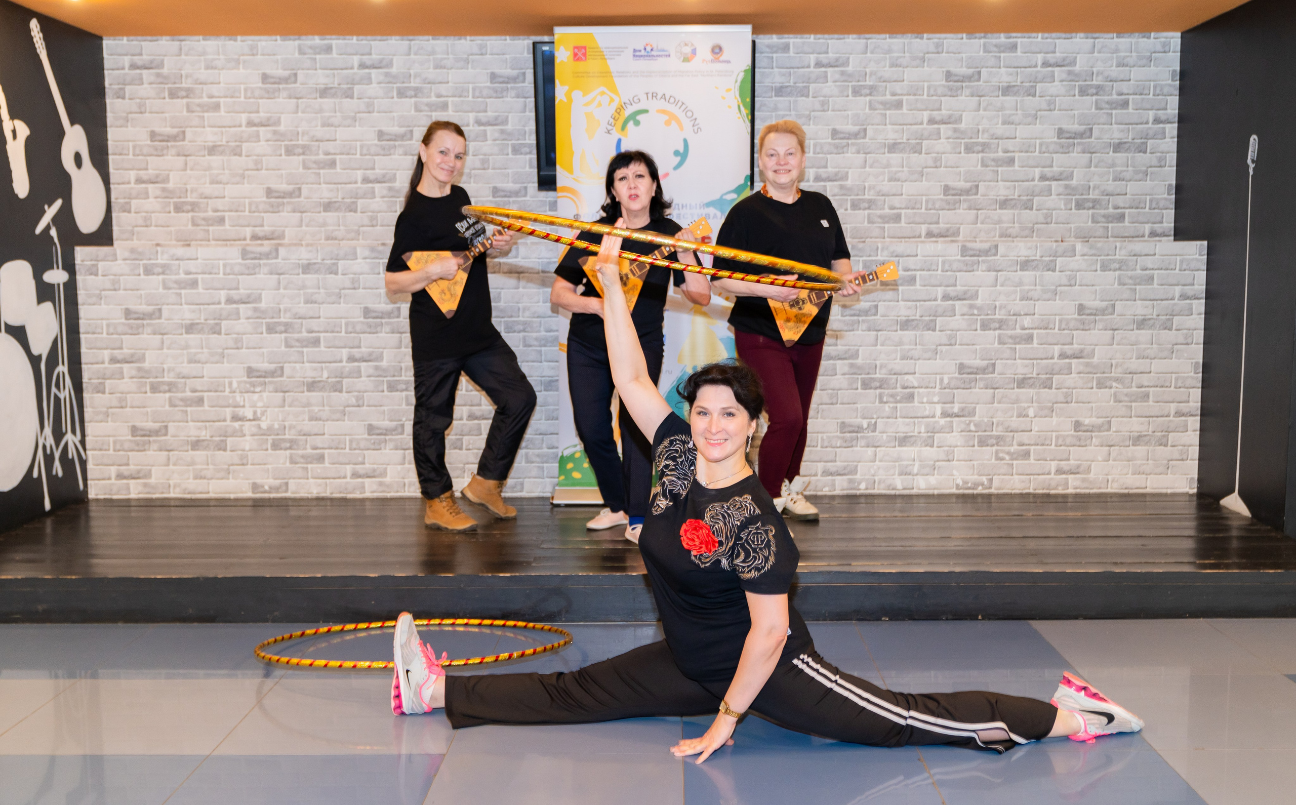 Участница XII Международного фольклорного фестиваля «Сохраняем традиции» Санкт-Петербург 2026 выполняет шпагат с обручами, за ней музыкантши с балалайками. Фото: Мальцев Евгений, evrednix.ru.