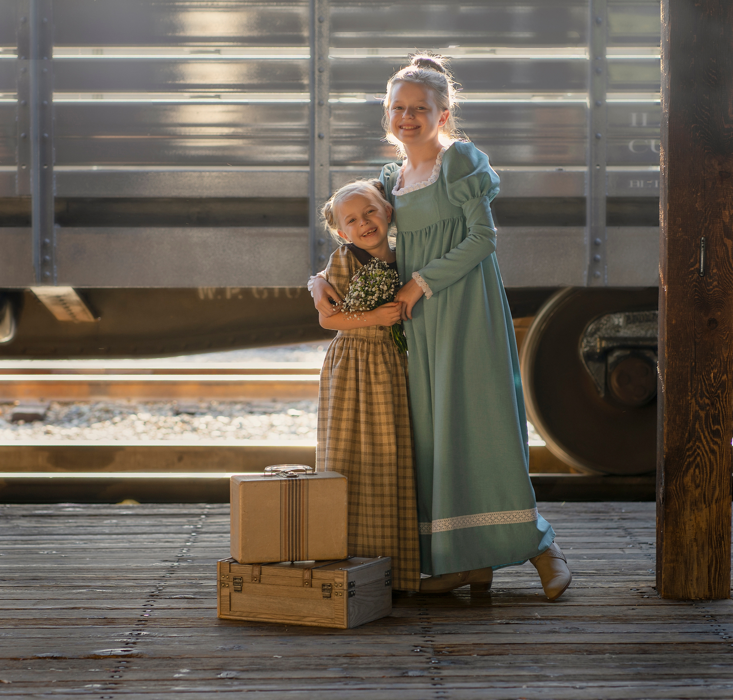 Две сестрички над чем-то смеются на перроне вокзала. На заднем плане стоит вагон.
