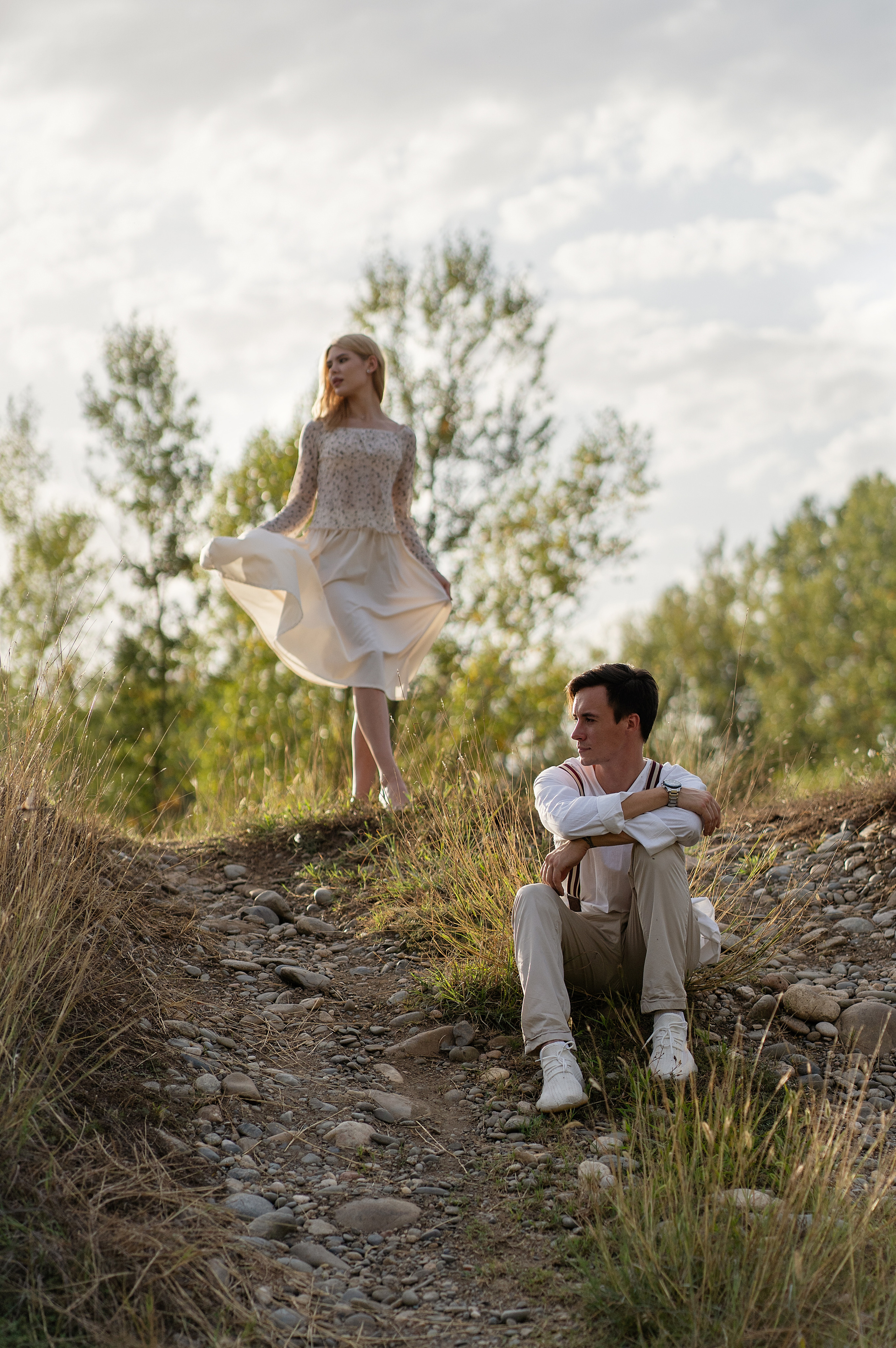 Парень сидит на пригорке, а девушка, чуть выше, слегка пританцовывает.