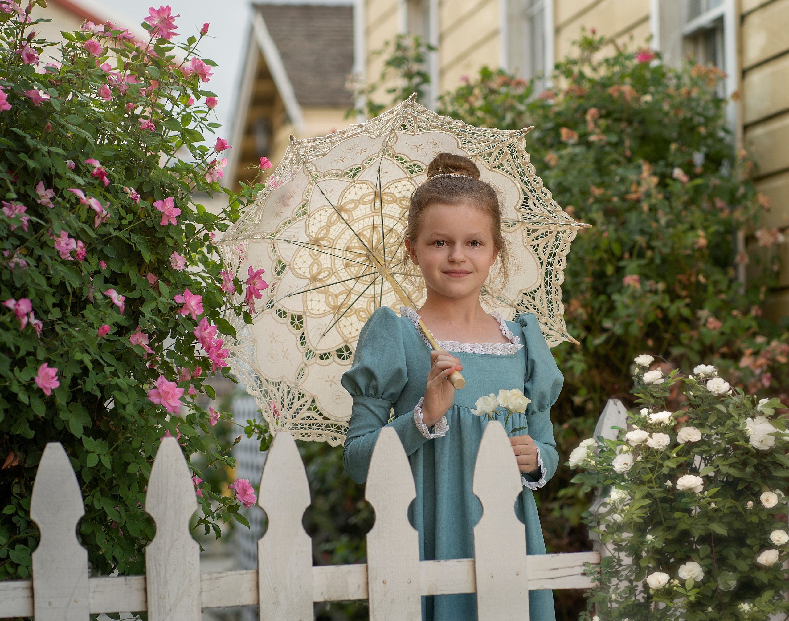 Девушка с кружевным зонтиком выглядывает через забор. Рядом растут розы.
