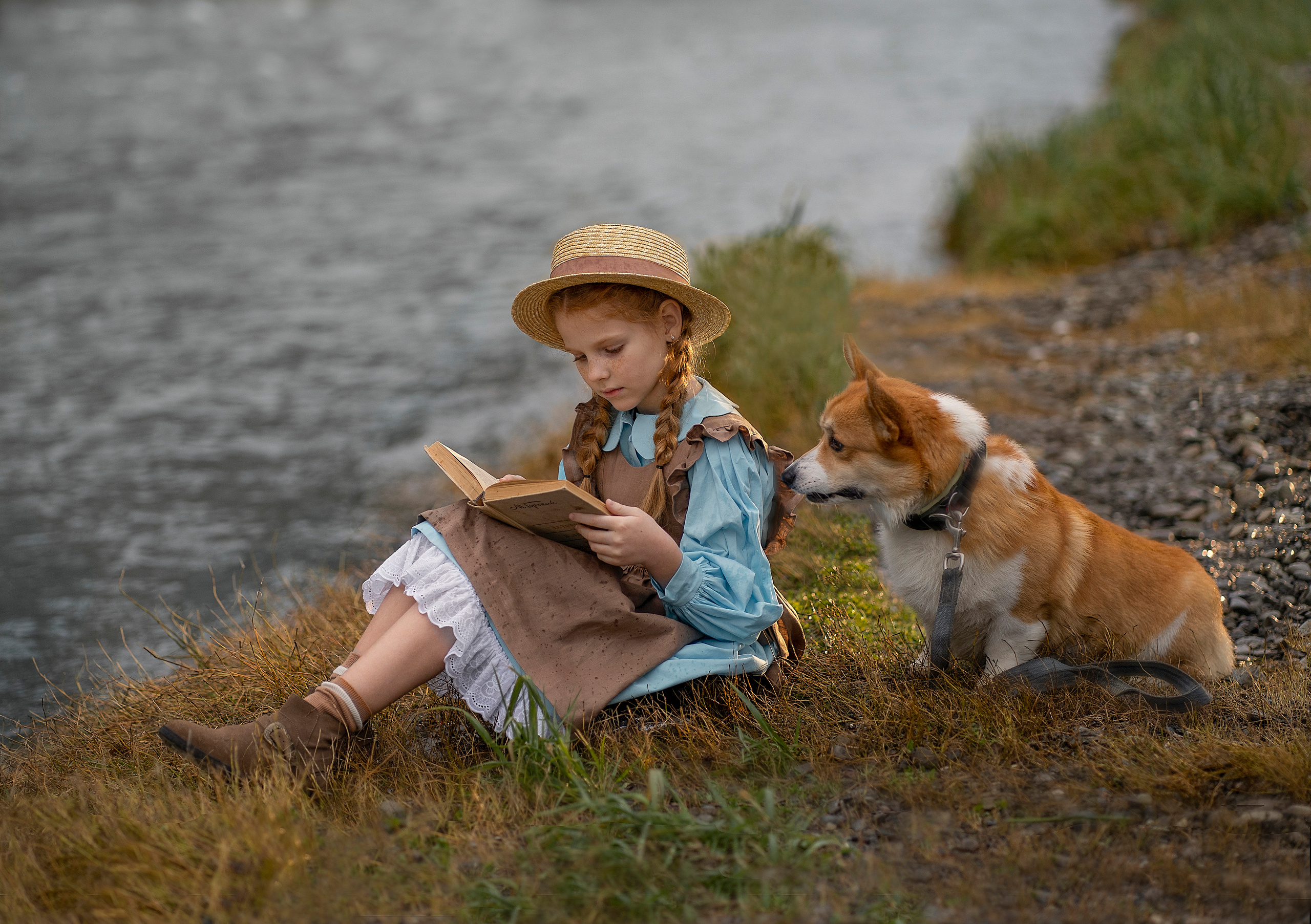 На берегу реки сидит девочка и читает книжка. Сзади подкралась собачка и заглядывает тоже в книжку.
