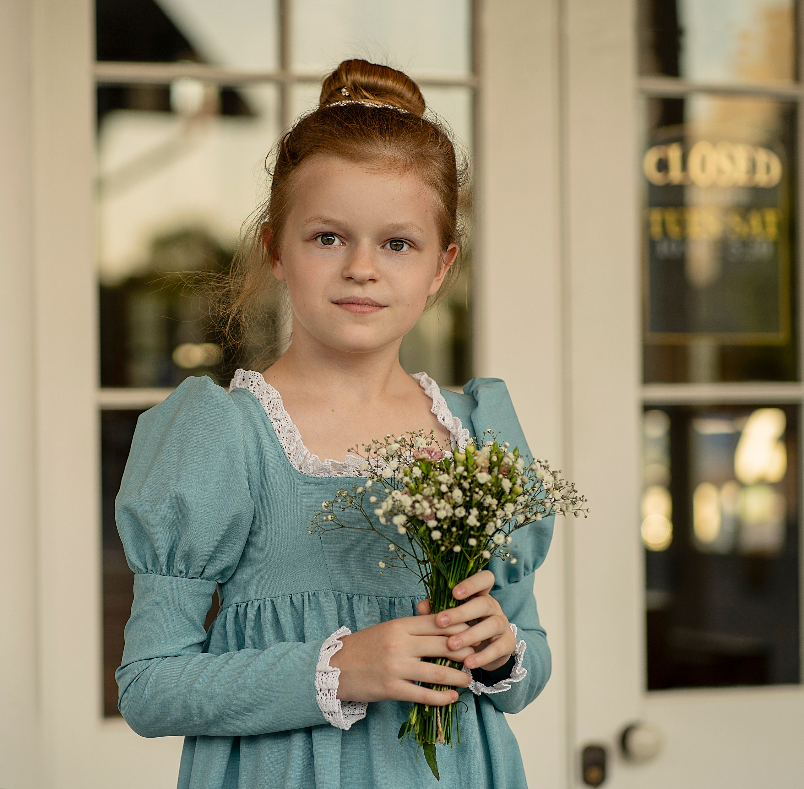 Девушка в бальном платье стоит на пороге дома, держит в руках букетик цветов.