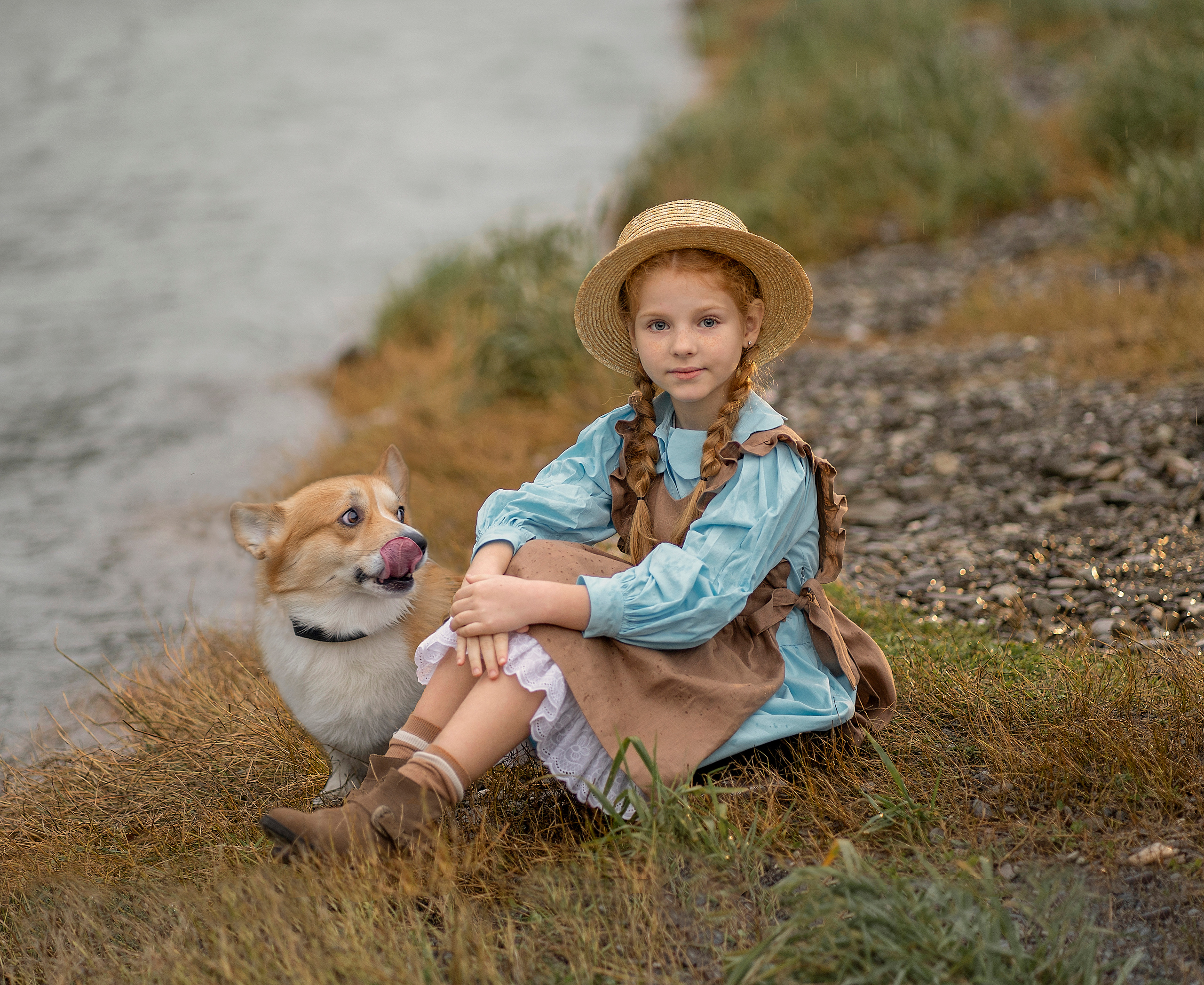 Девочка с рыжими косичками сидит на берегу речки. Рядом с ней облизывается собачка.