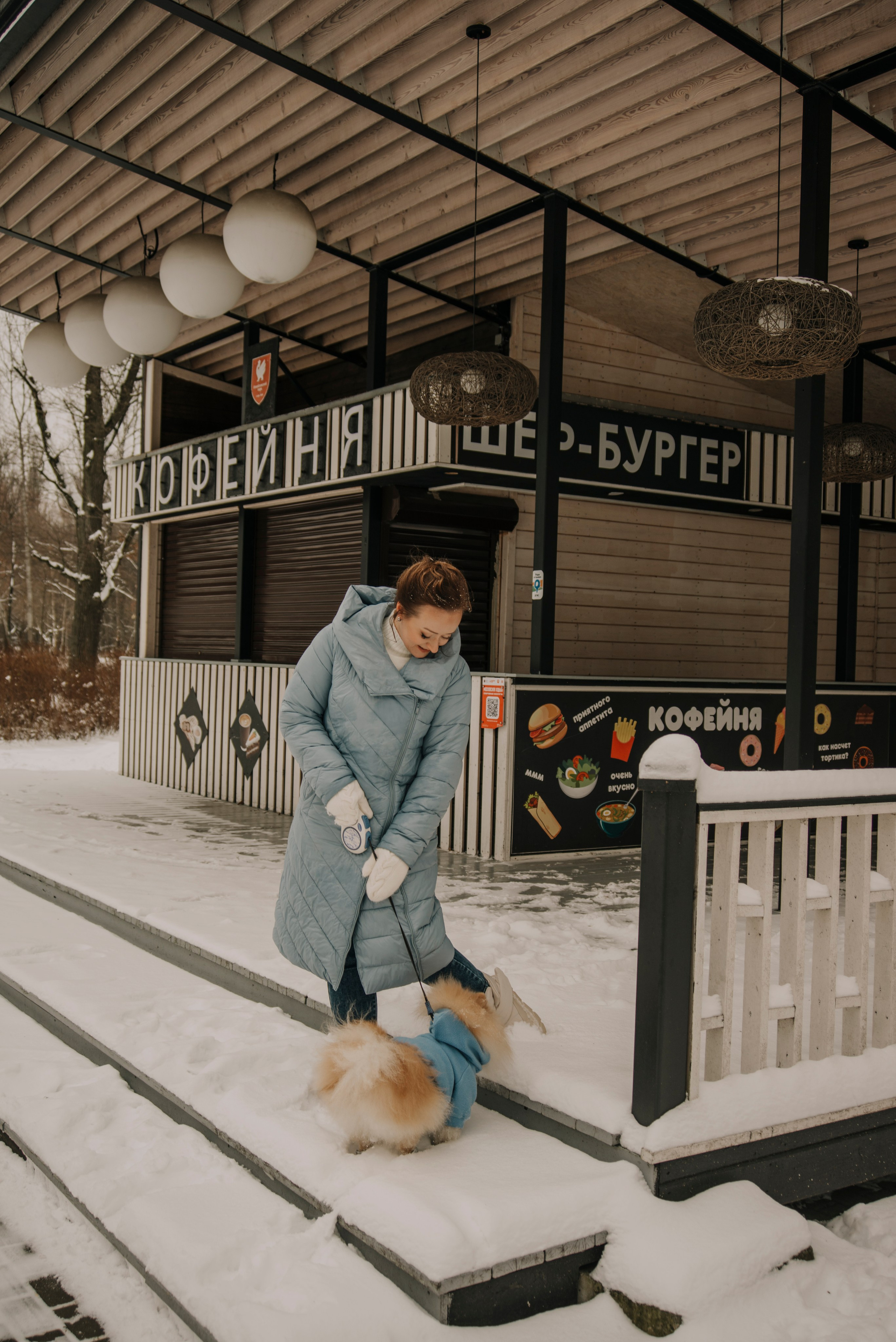 Фотография девушки в голубом пальто гуляющей по зимнему парку с собакой шпицем в голубом костюме
