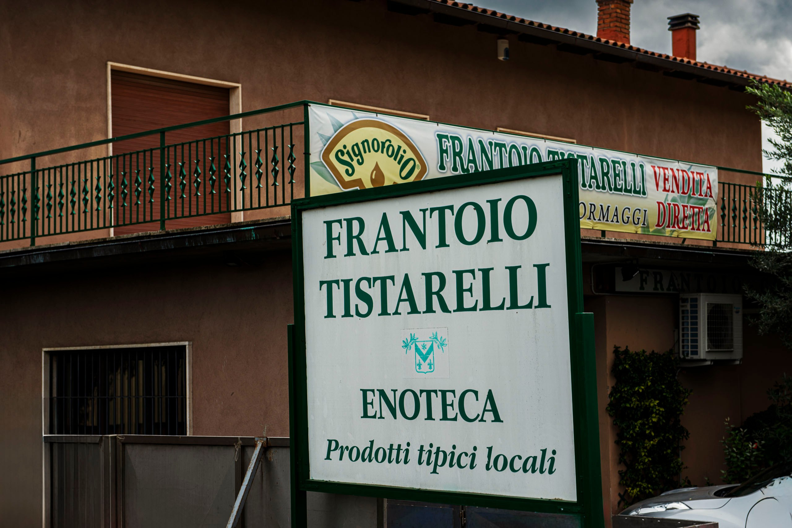 Вывеска с надписью "FRANTOIO TISTARELLI ENOTECA Prodotti tipici locali" перед зданием маслобойни с балконом и баннером на фоне.