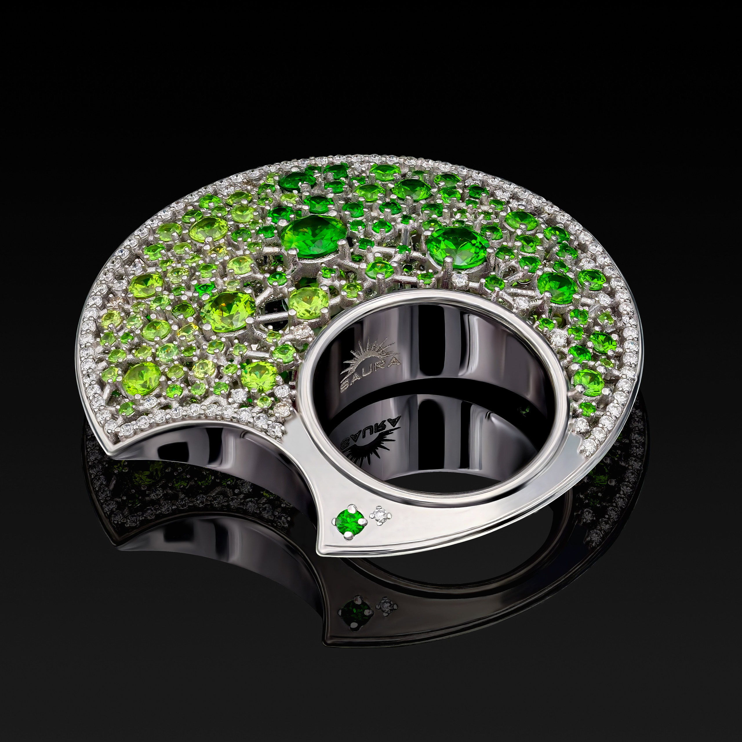 Круглое серебряное кольцо, украшенное множеством зеленых драгоценных камней и более мелкими прозрачными кристаллами на отражающей черной поверхности.