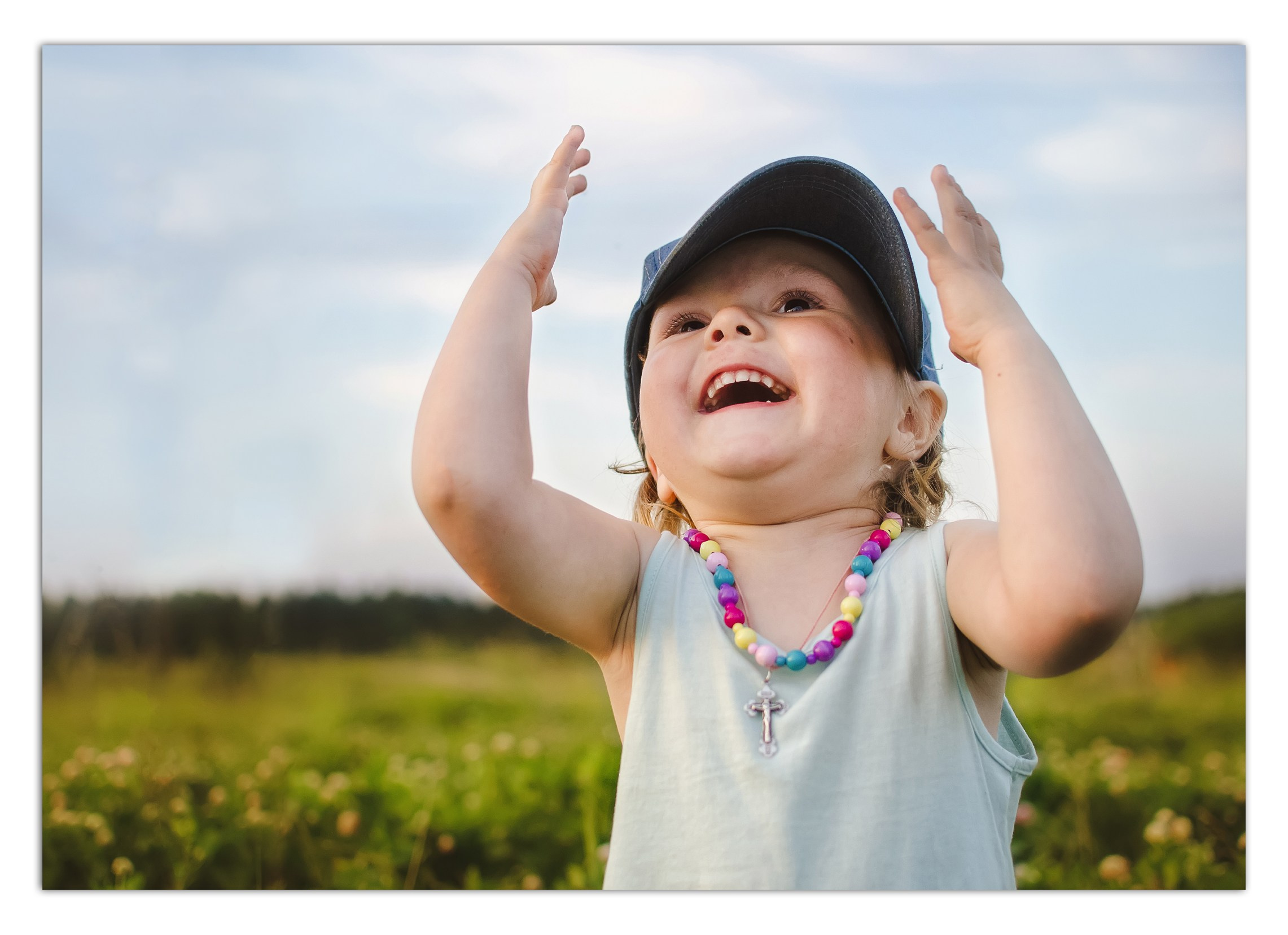 Детская фотосъемка на природе- девочка в кепке с яркими бусами играет на лугу.