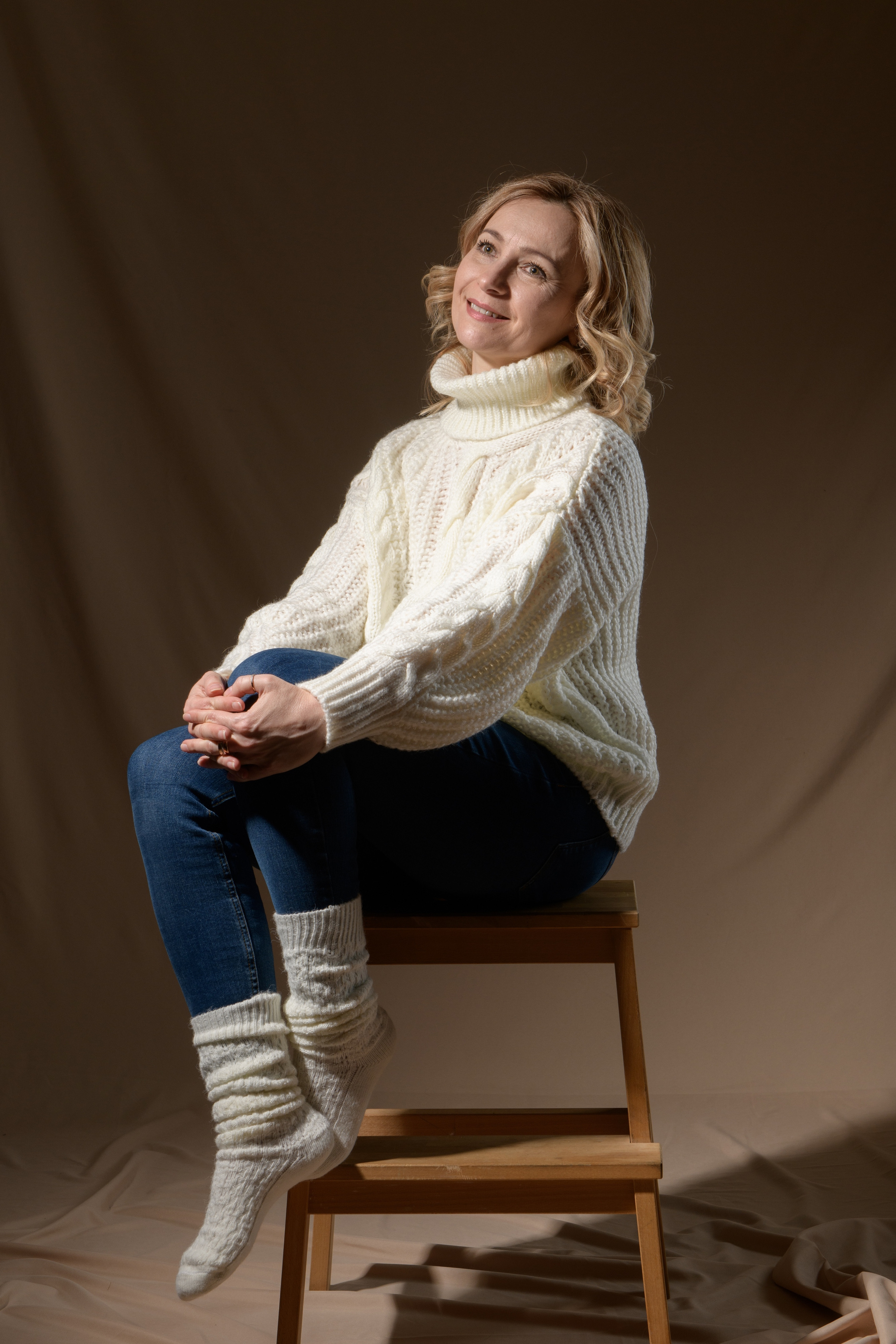 девушка в белом свитере и синих джинсах сидит на скамейке положив нагу на ногу и обняв колени руками