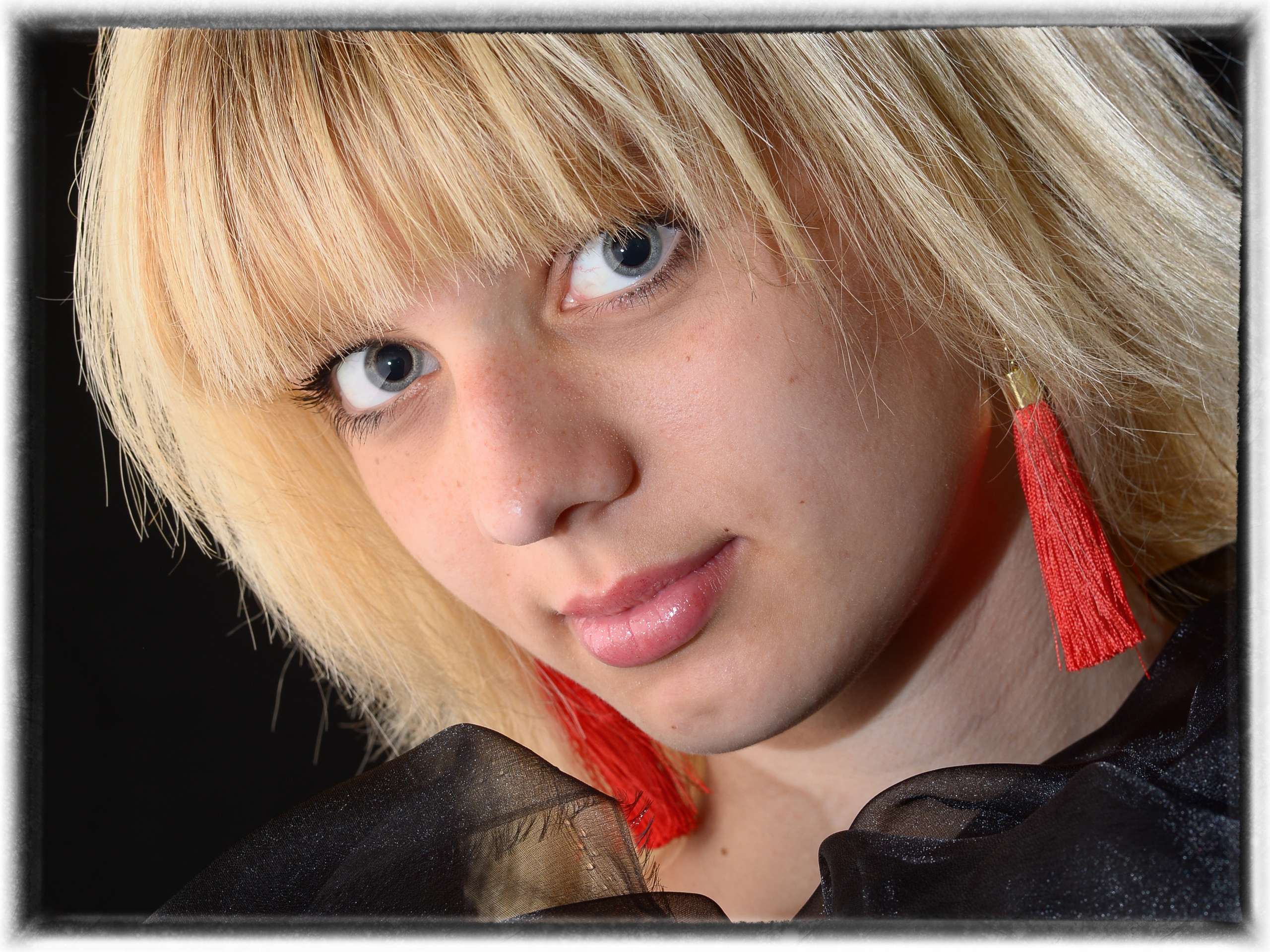 крупный портрет девушки со светлыми волосами, красными серьгами из ниточек и лукаво смотрящей в камеру фотоаппарата