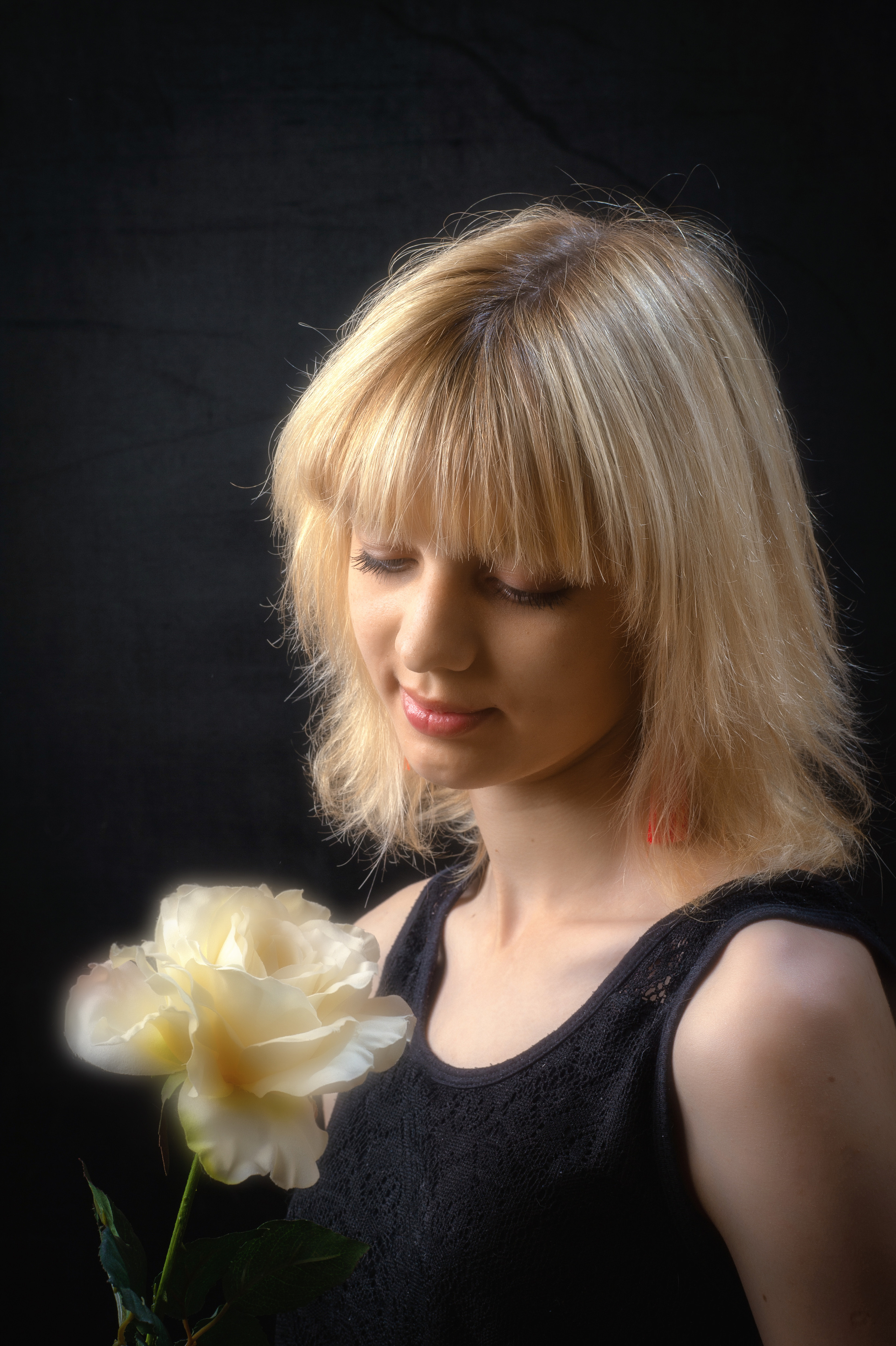 Портрет девушки с русыми волосами в черной майке  нежно смотрящей на цветок розы которую держит в руках