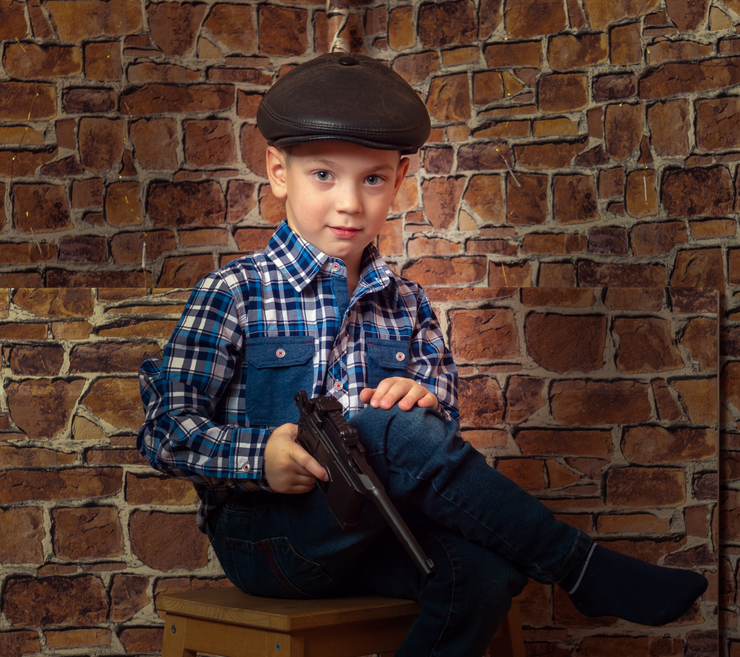 мальчик сидит на фоне кирпичной стены и держит пистолет маузер на голове у него кепка